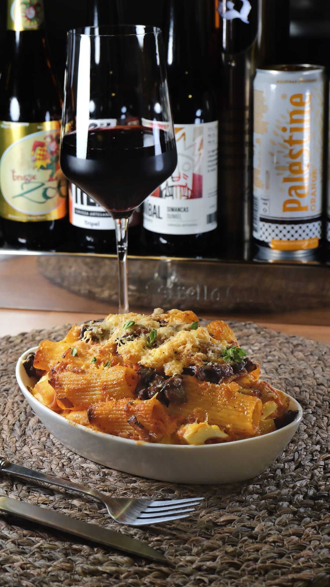 Un plato de pasta con salsa de tomate y hierbas, acompañado de una copa de vino tinto y botellas de vino.