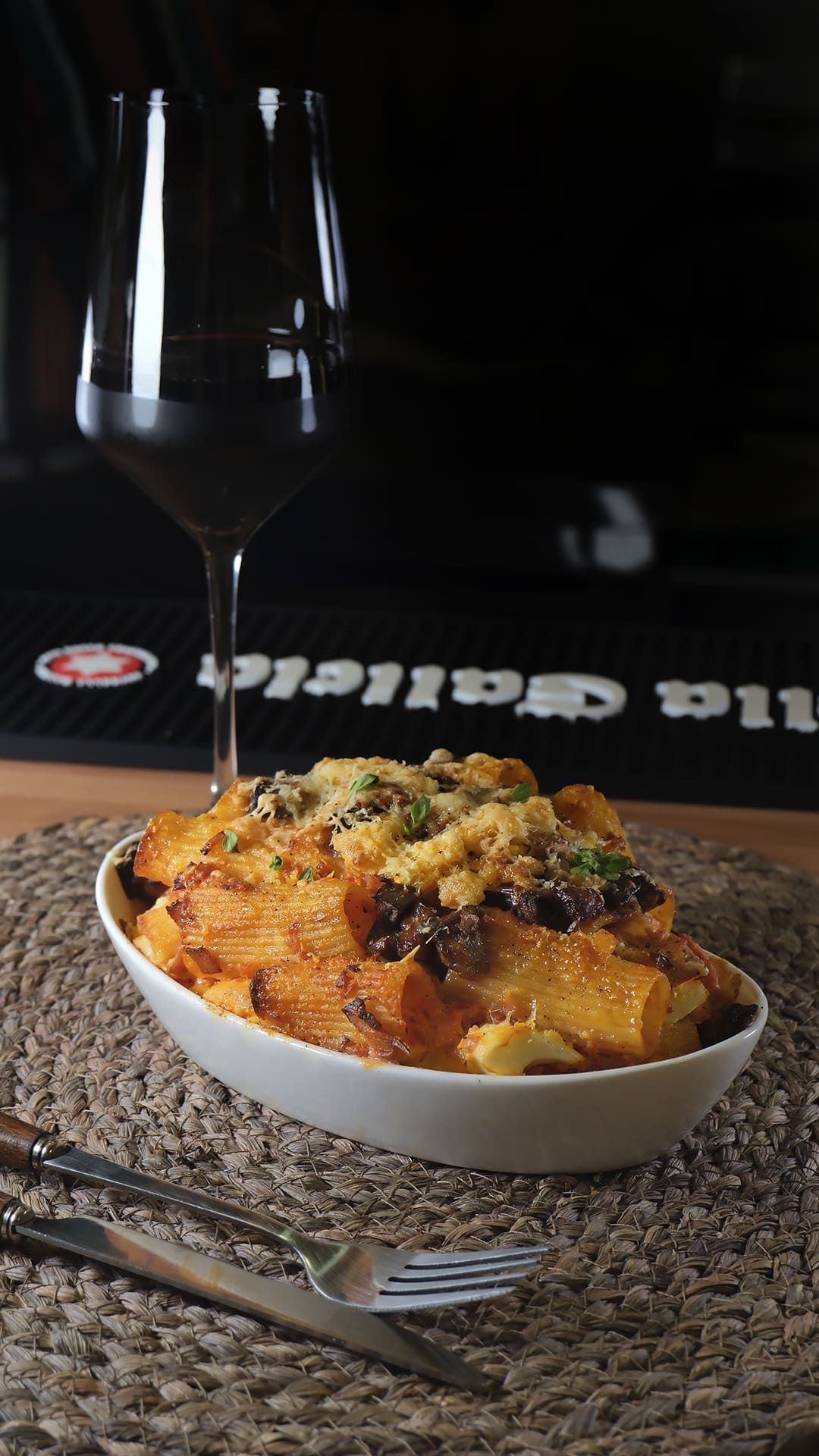 Un plato de pasta al horno con hierbas aromáticas, junto a una copa de vino sobre un mantel individual tejido, en una mesa oscura.