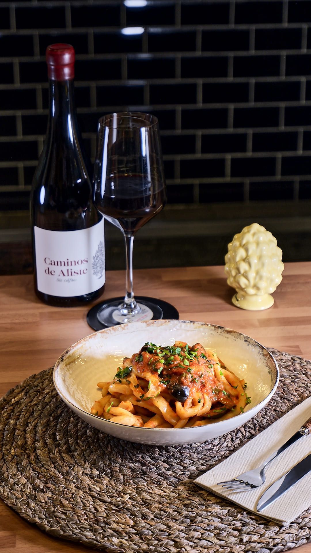 Plato de pasta con salsa roja, botella y copa de vino y piña decorativa sobre una mesa de madera.