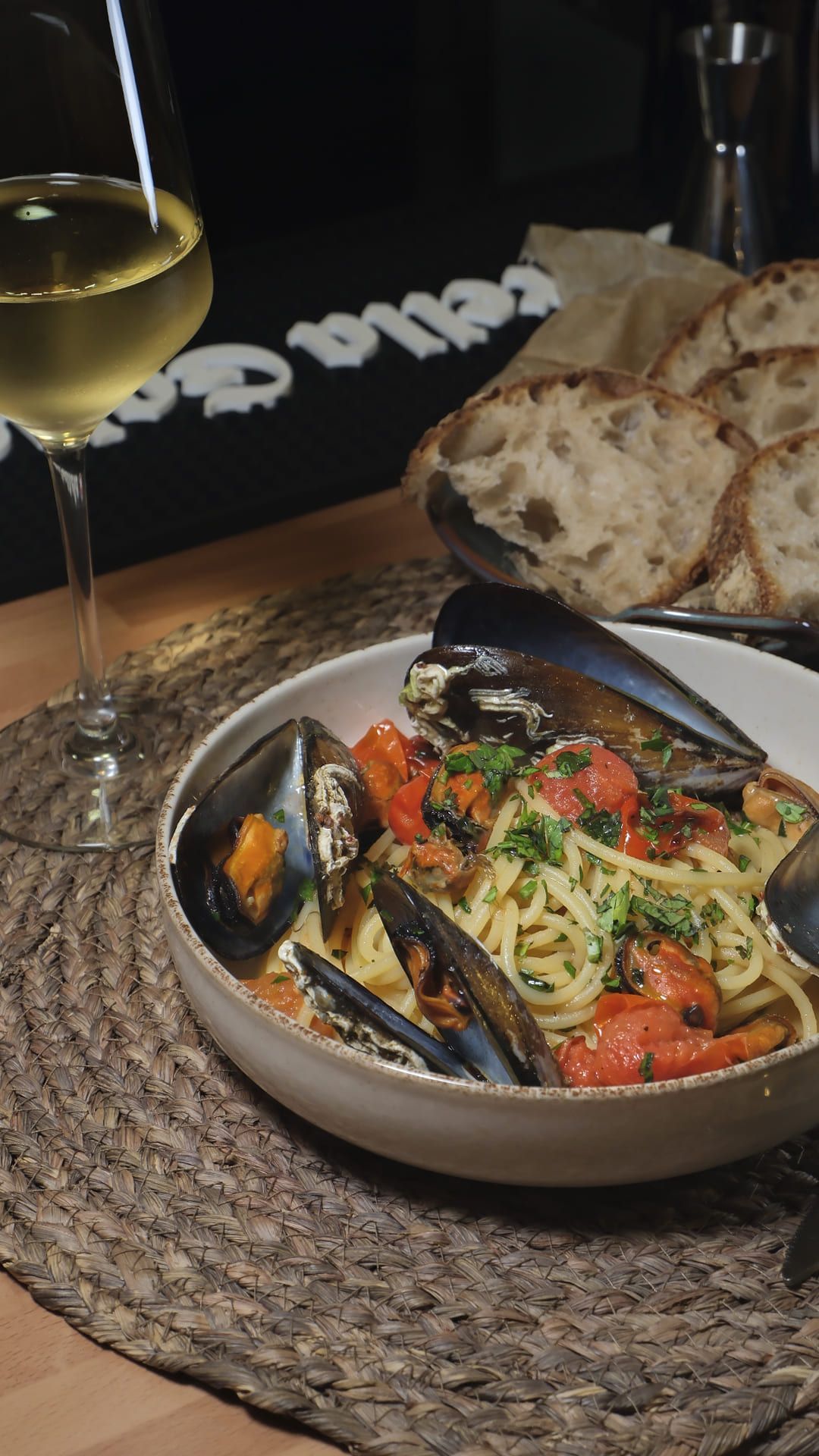 Mesa de comedor con un cuenco de mejillones y verduras, pan y una copa de vino blanco sobre un mantel individual tejido.