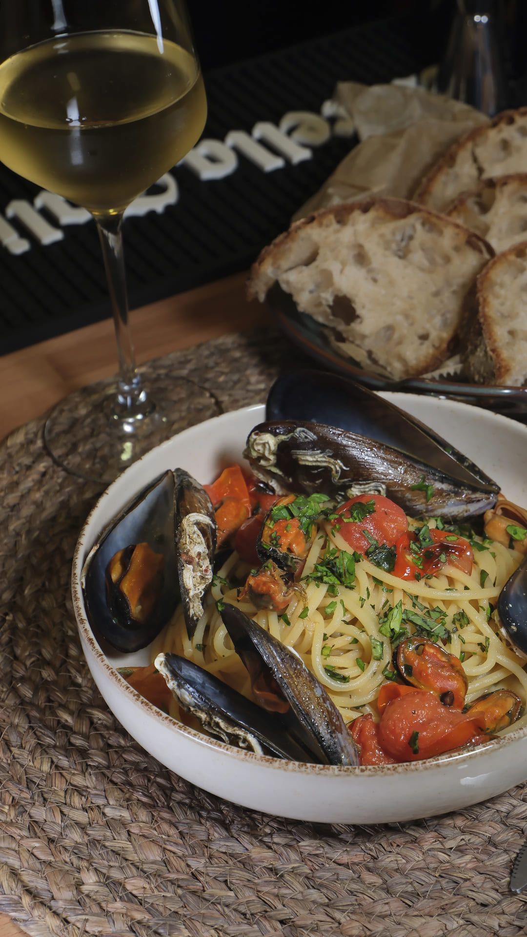 Pasta con mariscos, mejillones y tomates, acompañada de una copa de vino blanco y pan sobre una mesa.