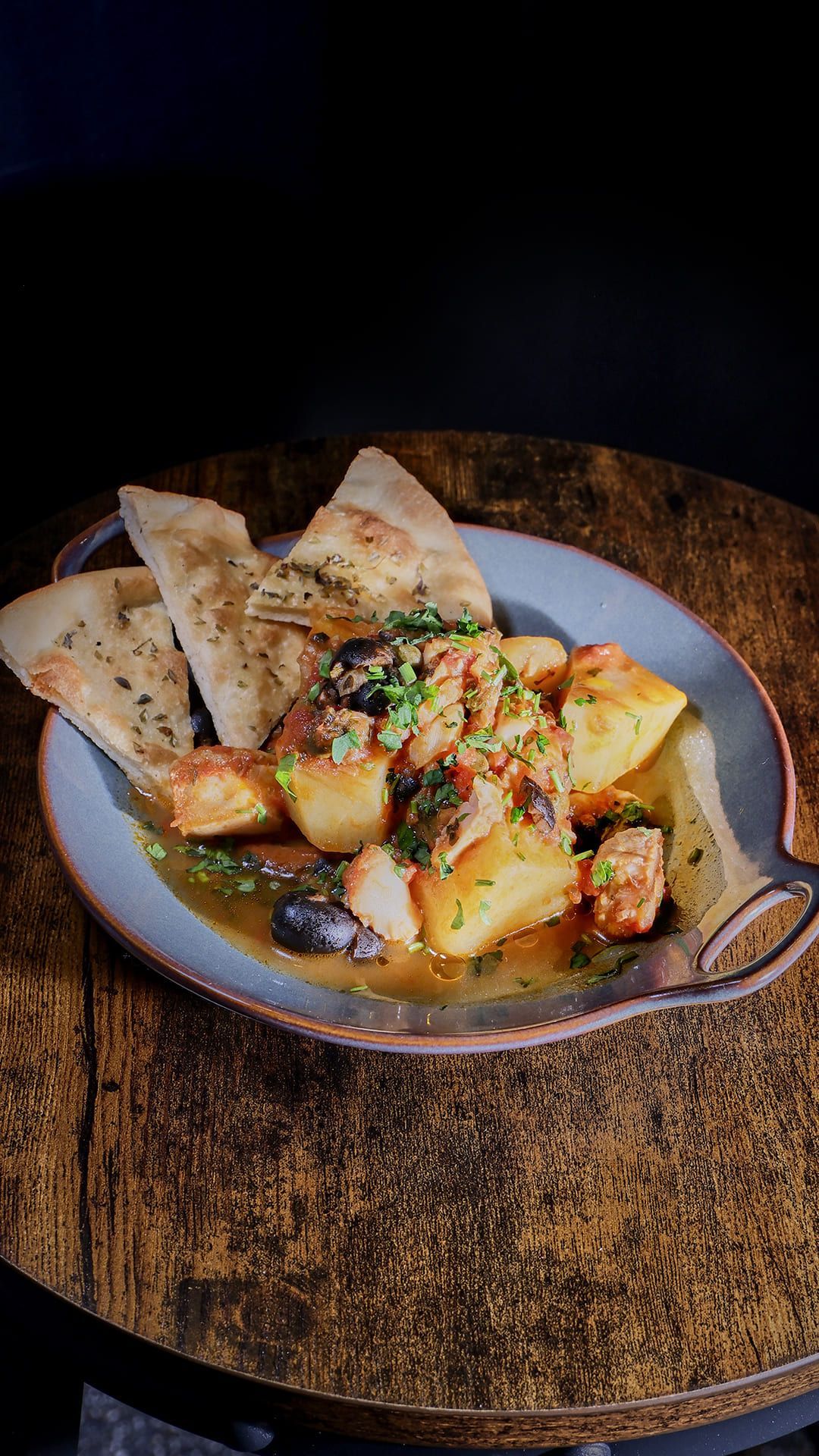Guiso con patatas, aceitunas y pan en un plato rústico, colocado sobre una mesa de madera.