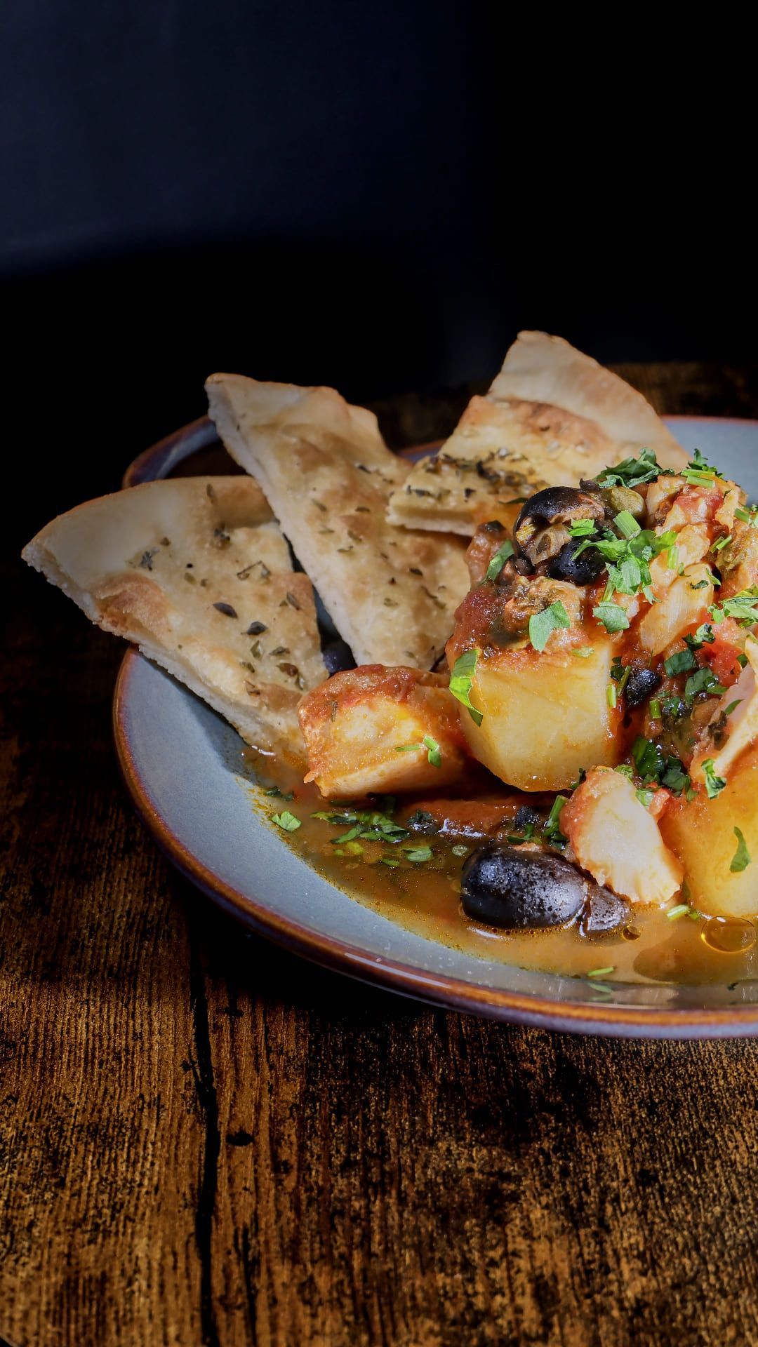 Primer plano de un plato con patatas guisadas, aceitunas, hierbas y pan pita sobre una mesa de madera.