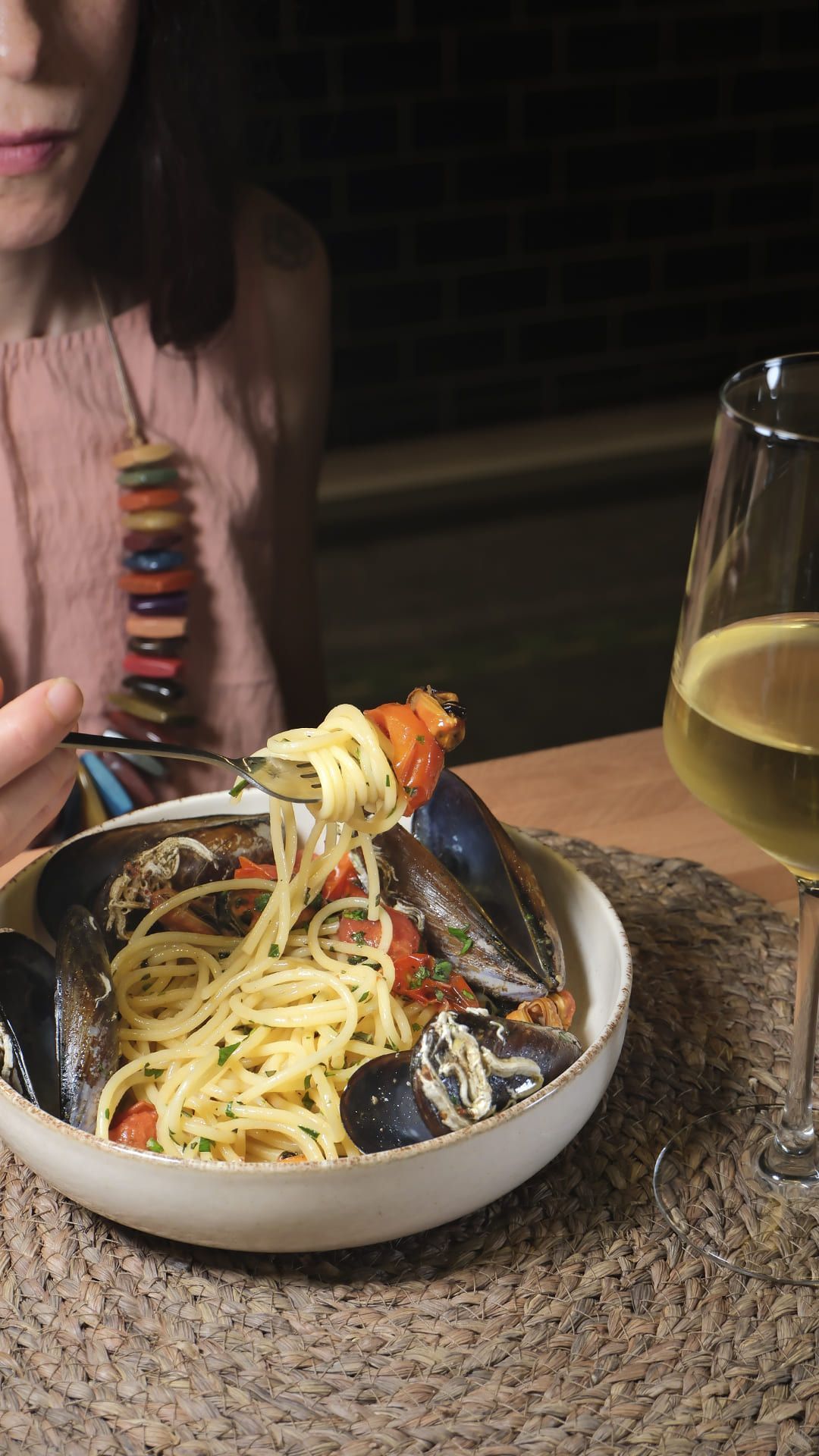 Una mujer come pasta con mejillones y bebe una copa de vino blanco en una mesa de un restaurante.