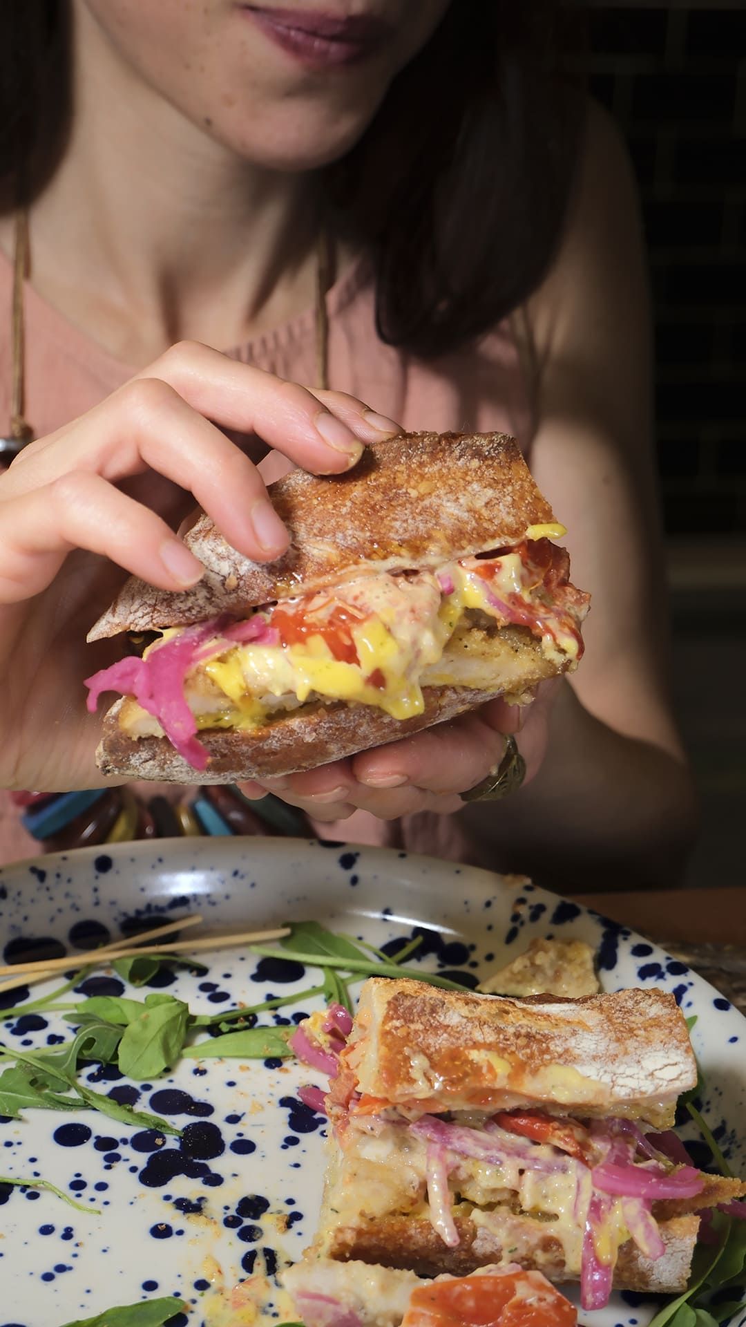 Una persona sostiene un sándwich desordenado sobre un plato, con otro sándwich a medio comer sobre una mesa.