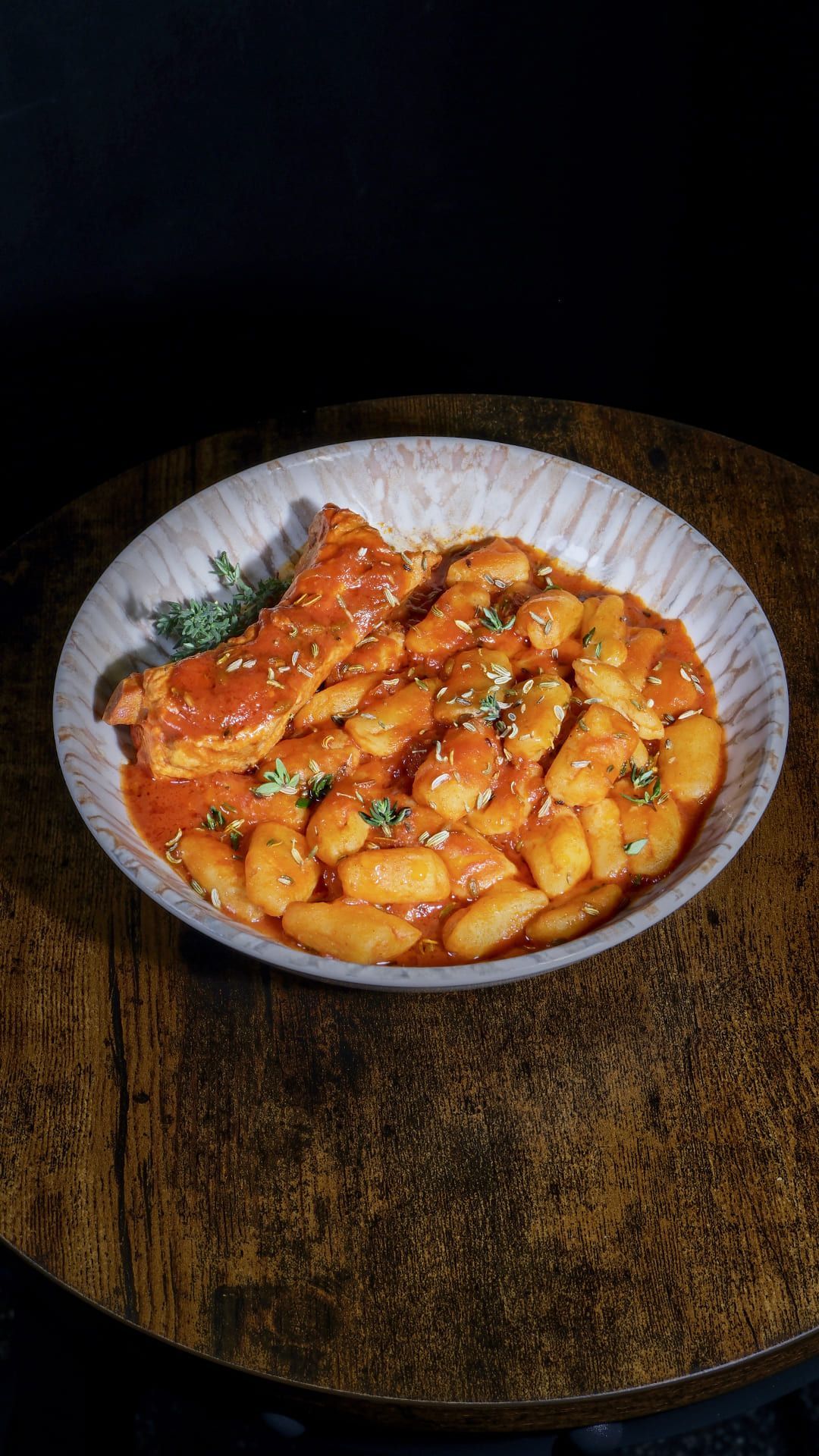 Tazón de tteokbokki (pasteles de arroz coreanos) en salsa roja, semillas de sésamo y guarnición de hierbas sobre una mesa de madera.