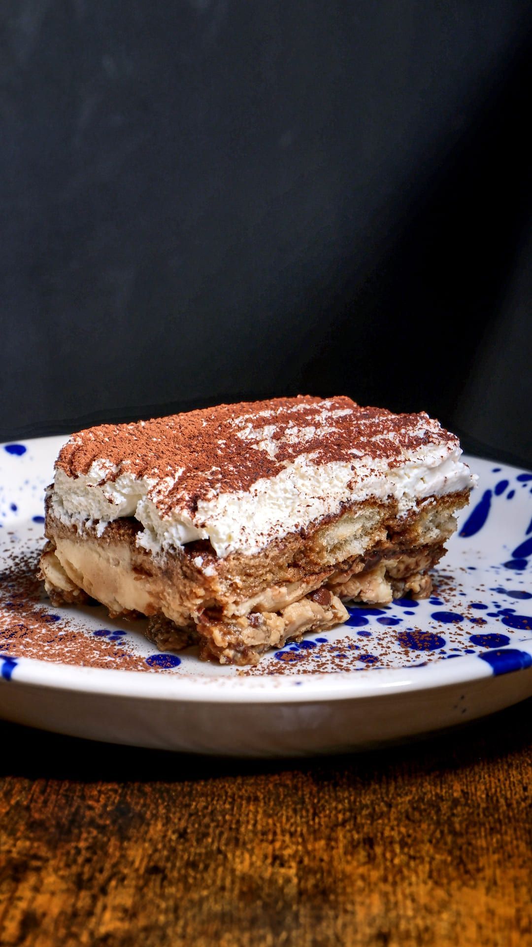 Rebanada de tiramisú espolvoreada con cacao en polvo sobre un plato moteado en blanco y azul.