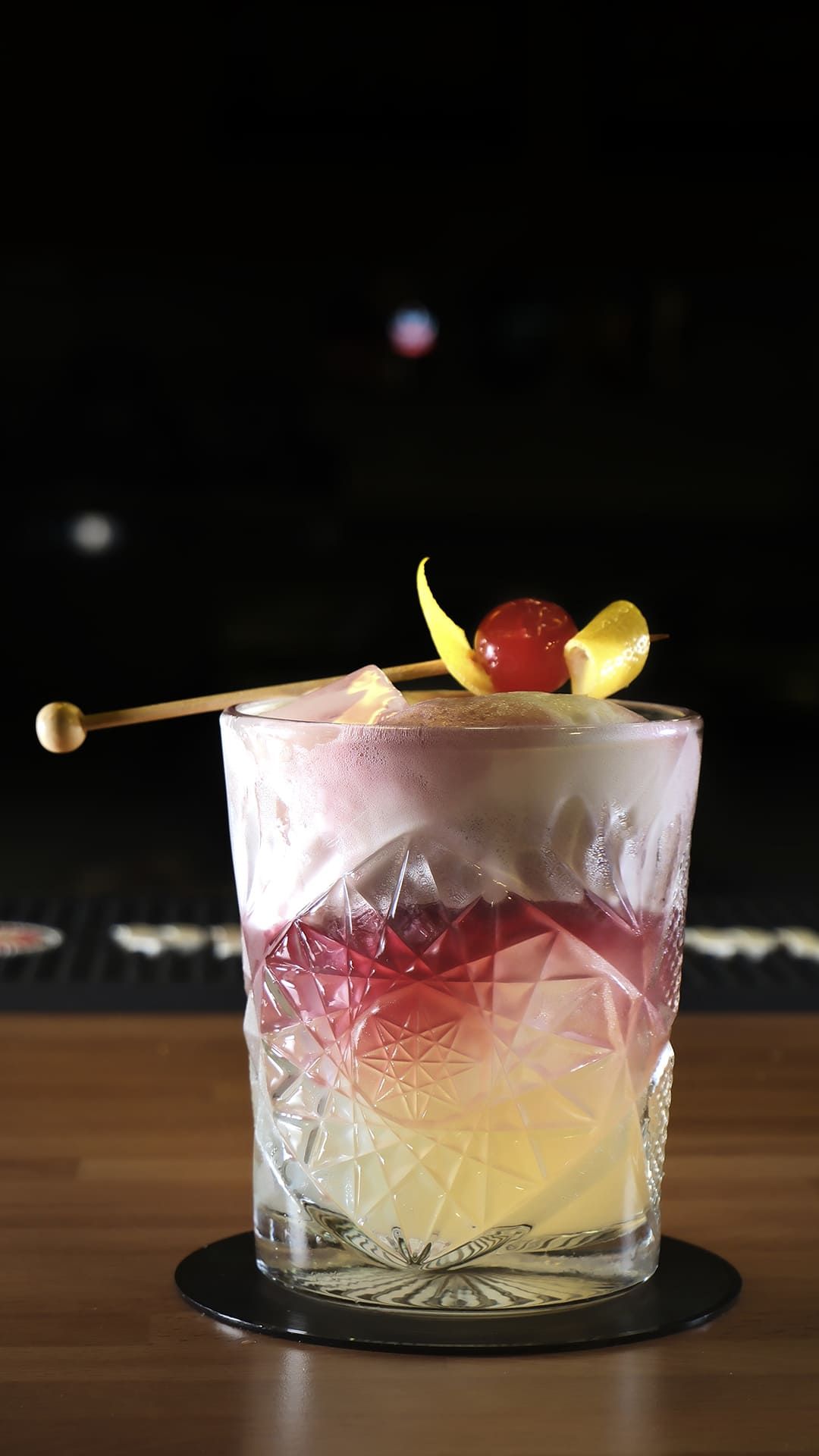 Cóctel en capas de color rosa y amarillo servido en un vaso escarchado, decorado con una cereza y rodajas de fruta.
