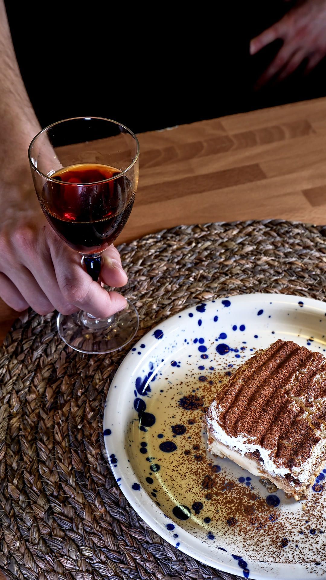 Persona sosteniendo un vaso de líquido oscuro, junto a un plato con postre espolvoreado con cacao, sobre una estera tejida.