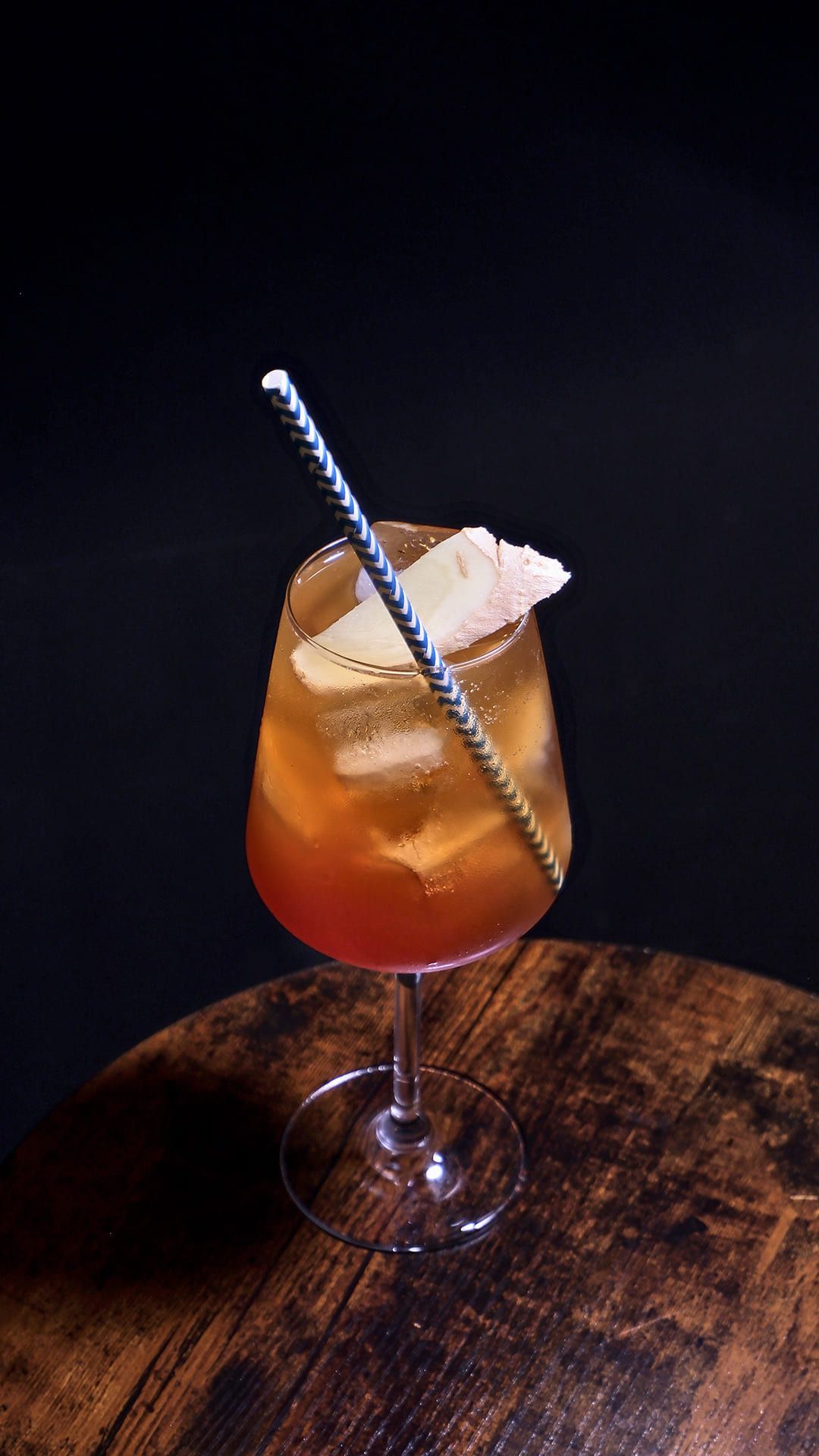 Cóctel en copa de tallo con hielo, pajita y guarnición, sobre una mesa de madera, sobre un fondo oscuro.