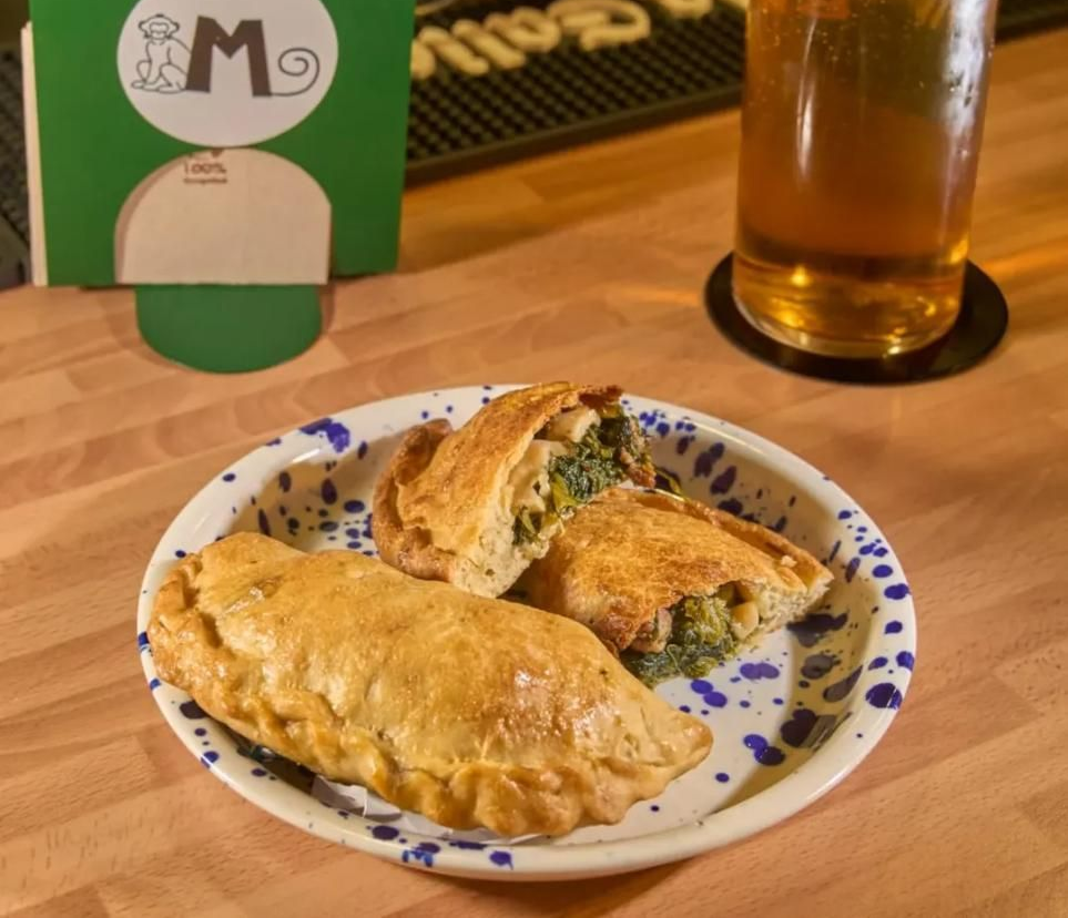 Dos pasteles dorados y rellenos en un plato, con una cerveza y un cartel verde en el fondo.