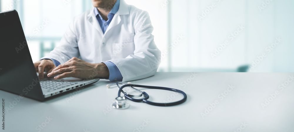 Medico che indossa un camice bianco mentre scrive su un computer portatile con uno stetoscopio sul tavolo.