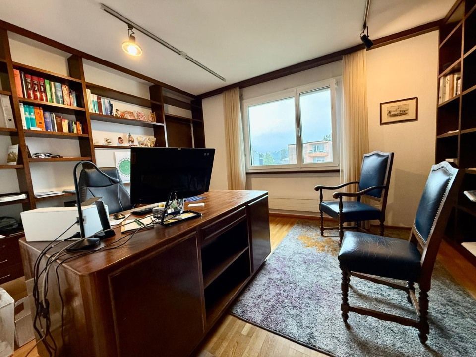 Ufficio con scrivania in legno scuro, librerie, due sedie blu e finestra.