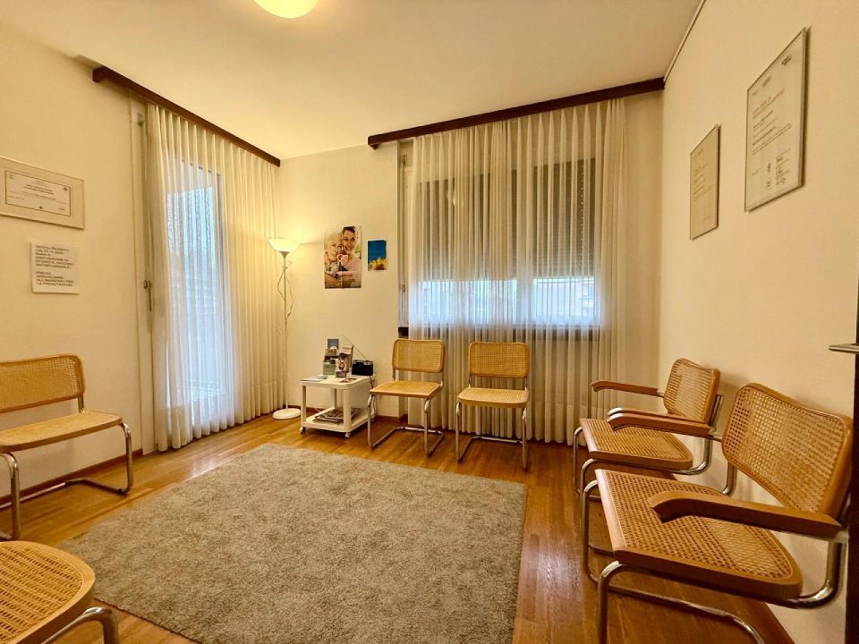 Sala d'attesa con sedie in legno, tappeto e finestre con tende.