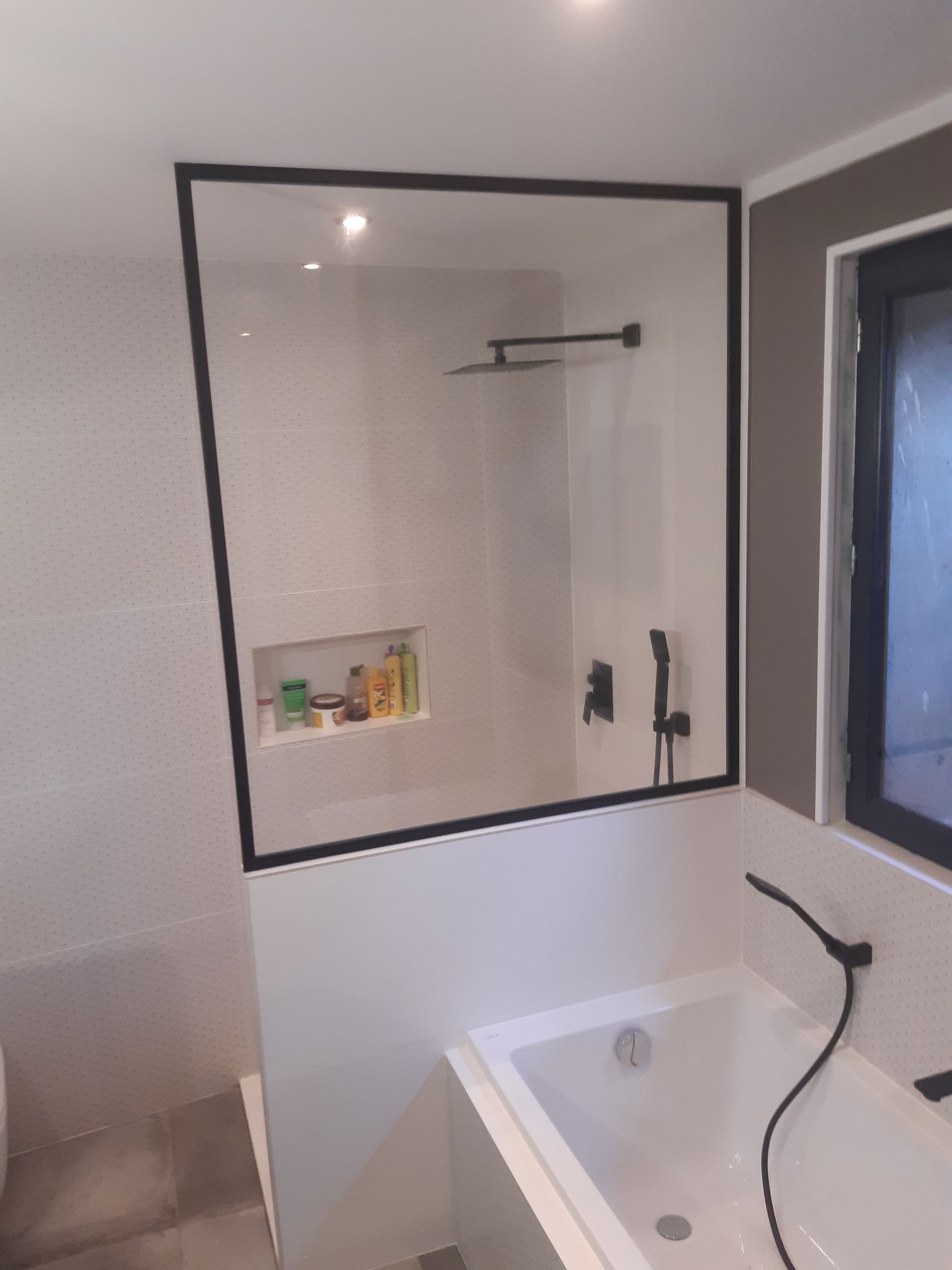 Salle de bains avec paroi de douche à cadre noir, baignoire et robinetterie noire sur carrelage blanc.
