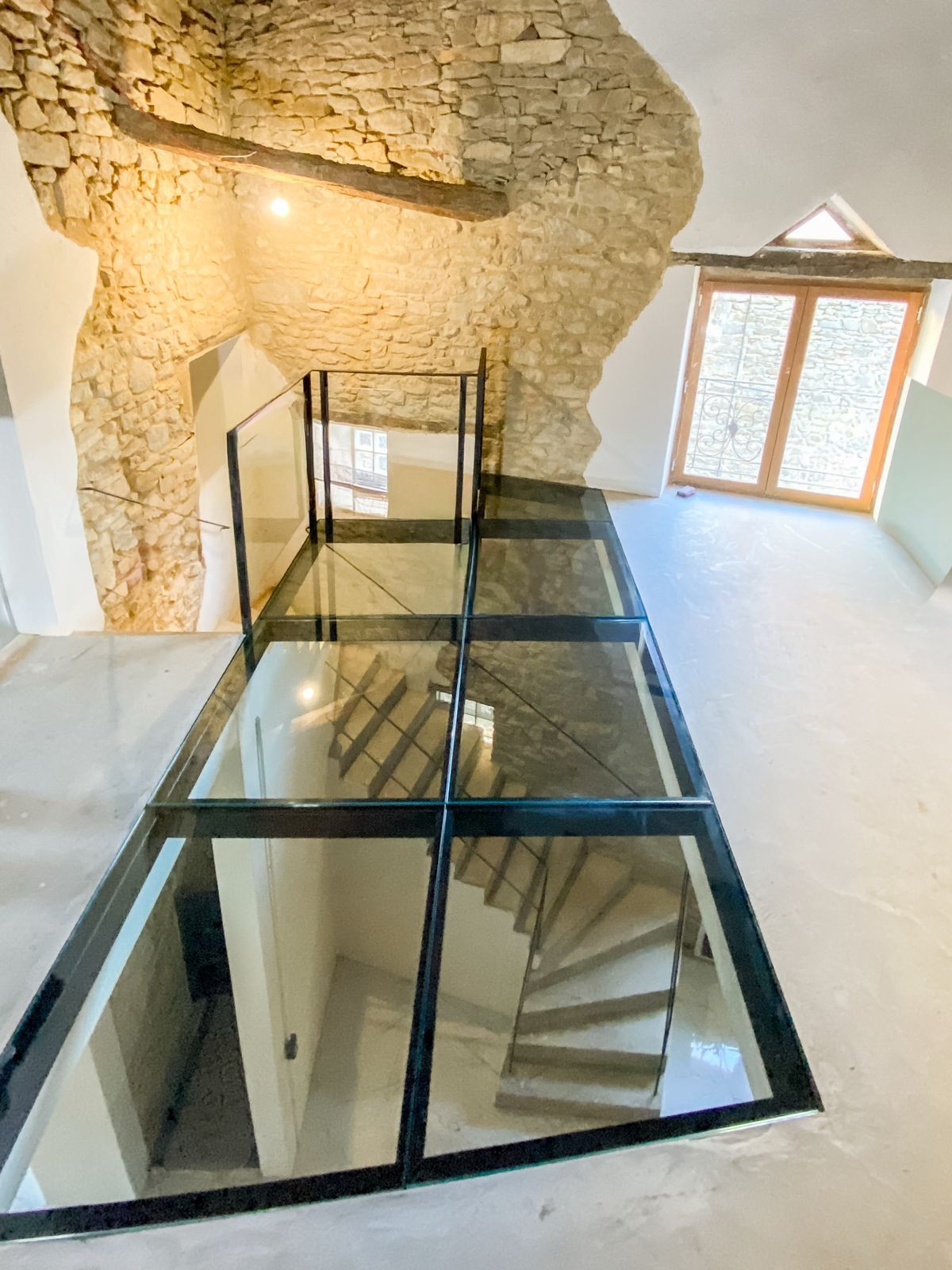 Passerelle vitrée surplombant un escalier dans un bâtiment aux murs de pierre, avec des portes à ossature bois et lumière naturelle.
