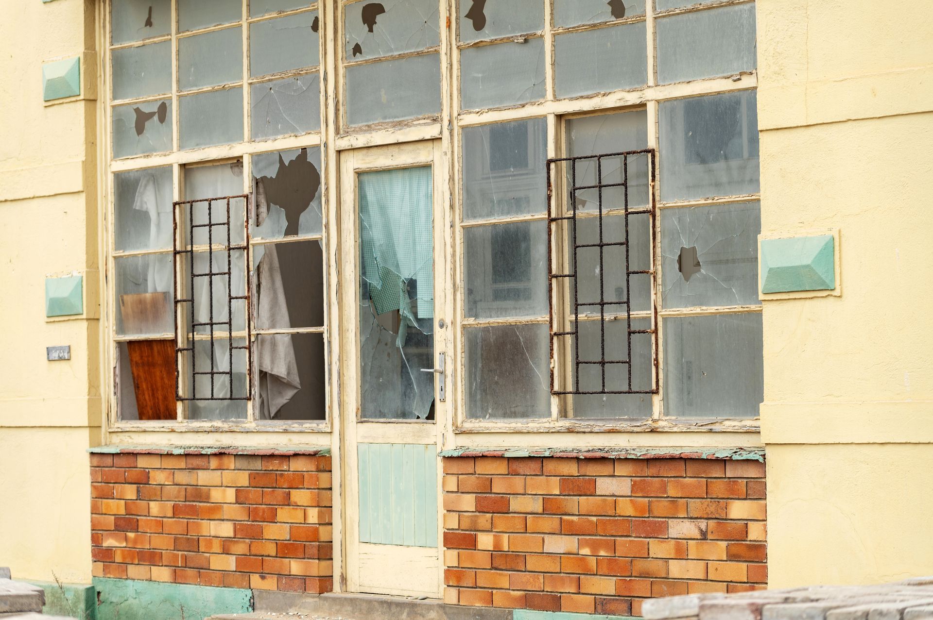 Façade d'immeuble délabrée avec fenêtres brisées, soubassement en briques et peinture jaune délavée.