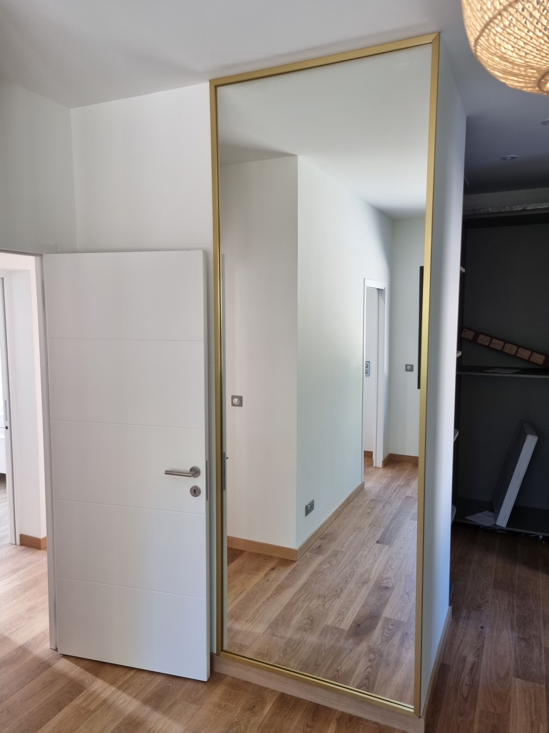 Un miroir en pied à cadre doré orne le couloir, reflétant l'espace. À gauche, une porte blanche et un parquet.