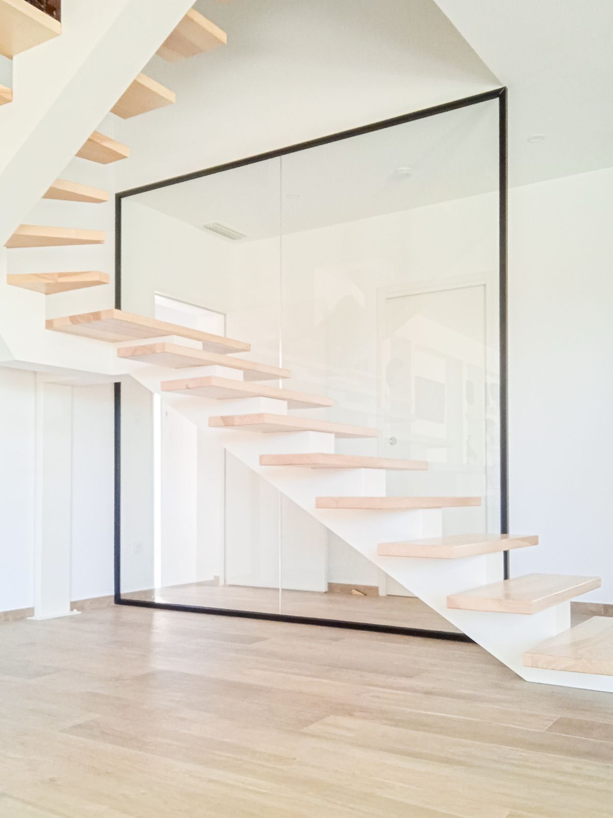 Escalier moderne avec marches en bois et panneau de verre encadré de noir, sur fond de murs blancs.