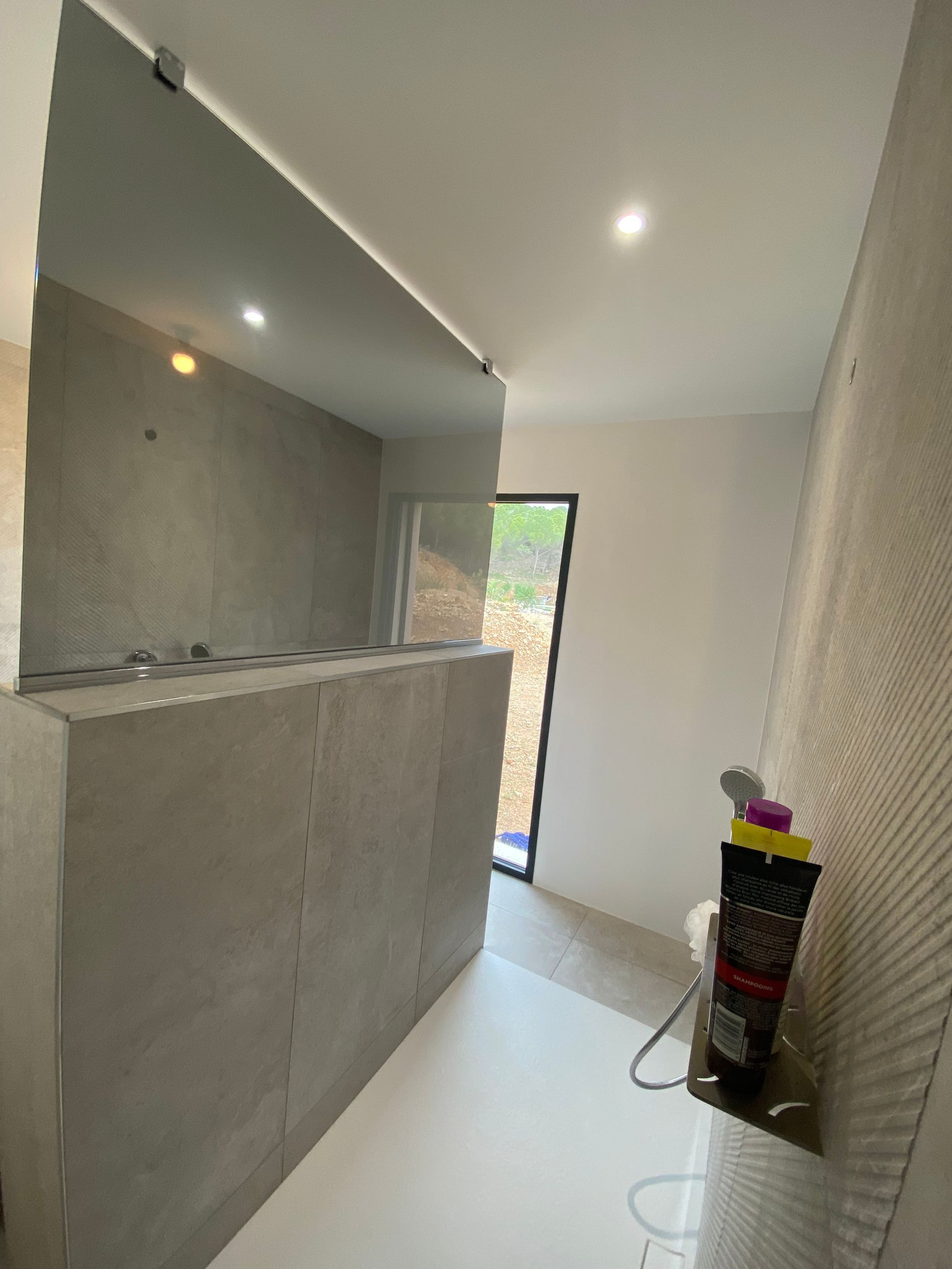 Salle de bains avec murs carrelés gris, douche et fenêtre donnant sur l'extérieur.