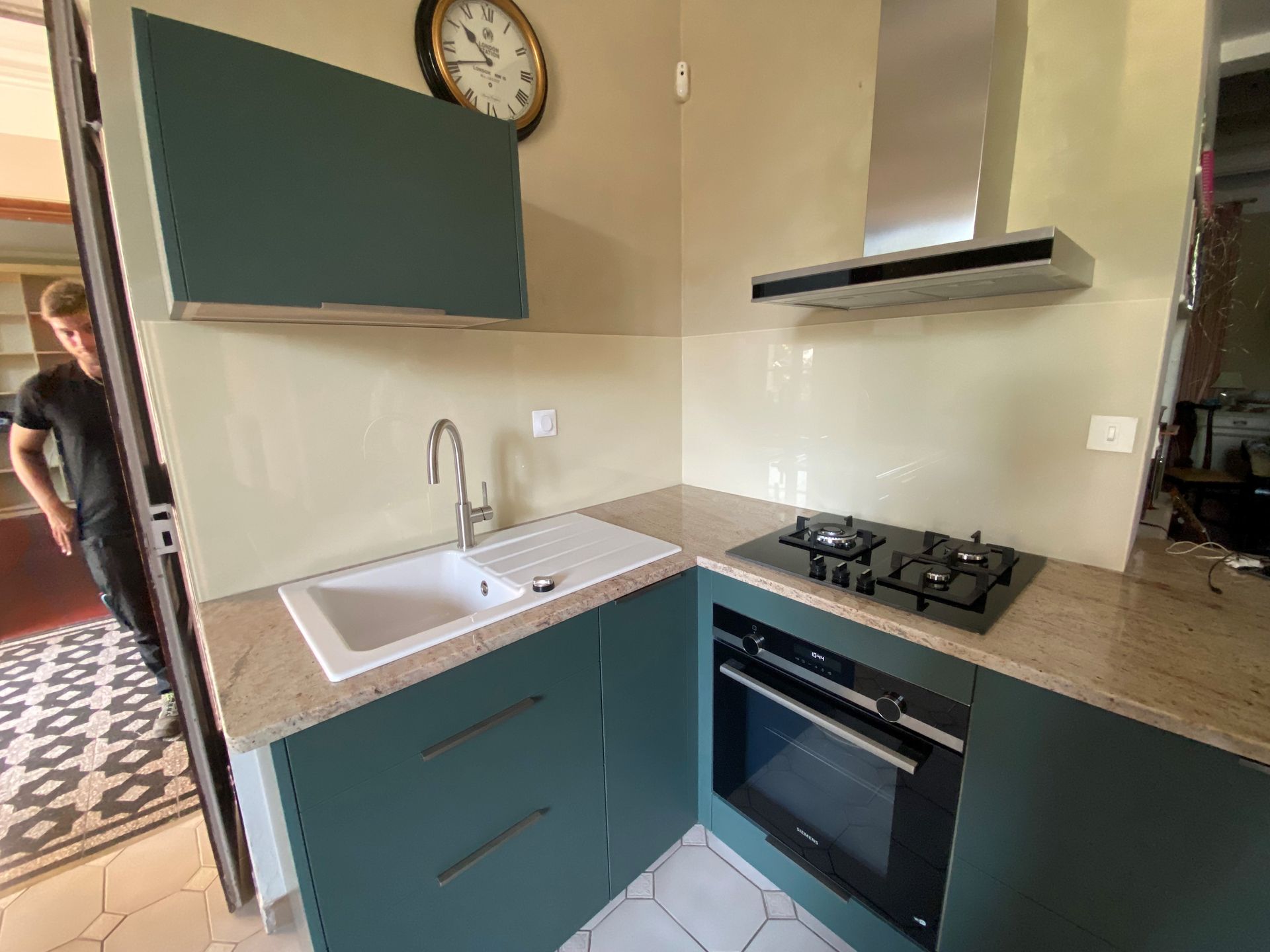 Petite cuisine turquoise avec évier, plaque de cuisson, four et hotte. Plan de travail et crédence beiges. Une personne se tient dans l'embrasure de la porte.