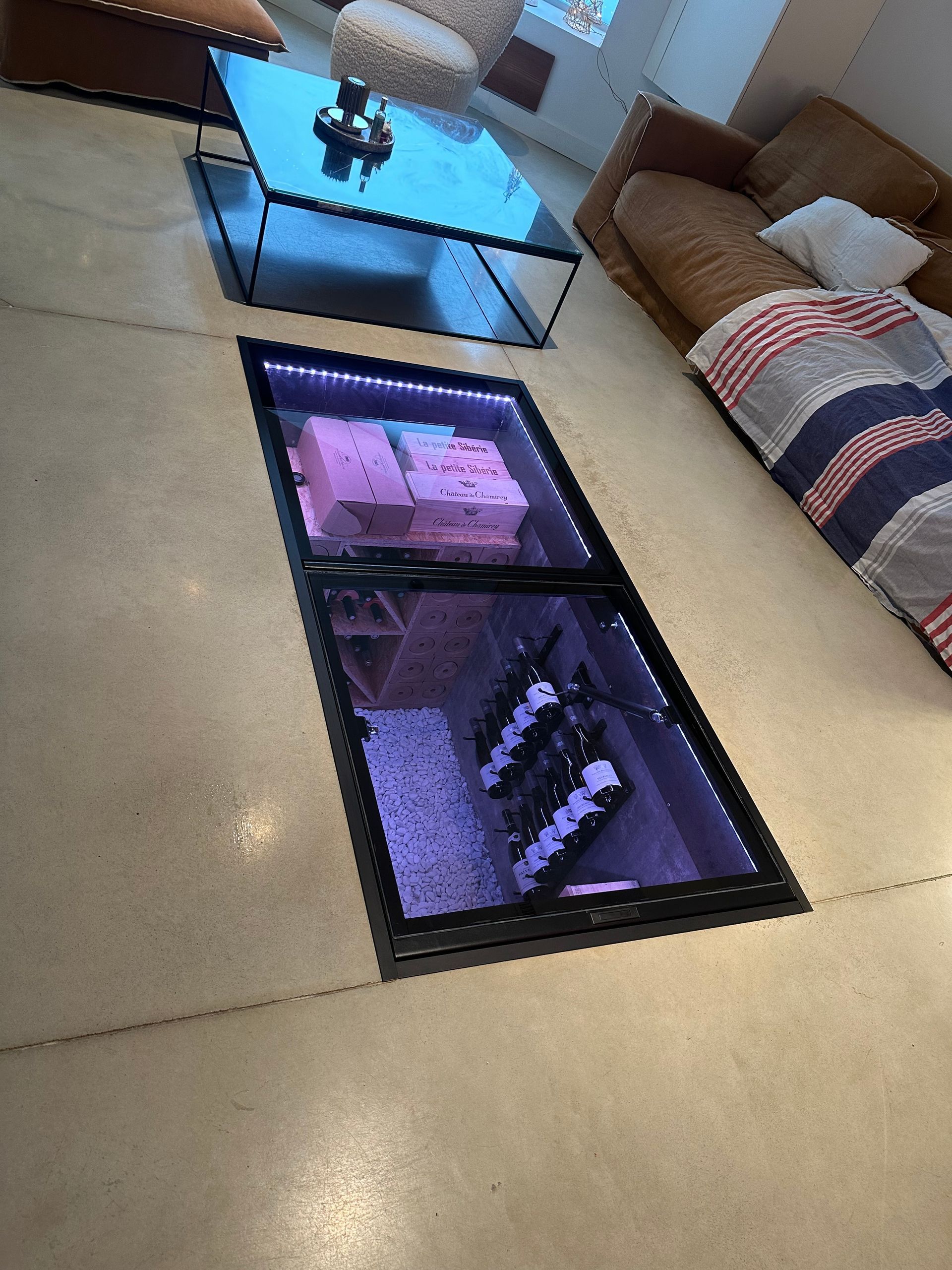 Cave à vin vitrée au sol dans un salon moderne, bouteilles visibles. Éclairage LED violet, sol aux tons neutres, canapé.