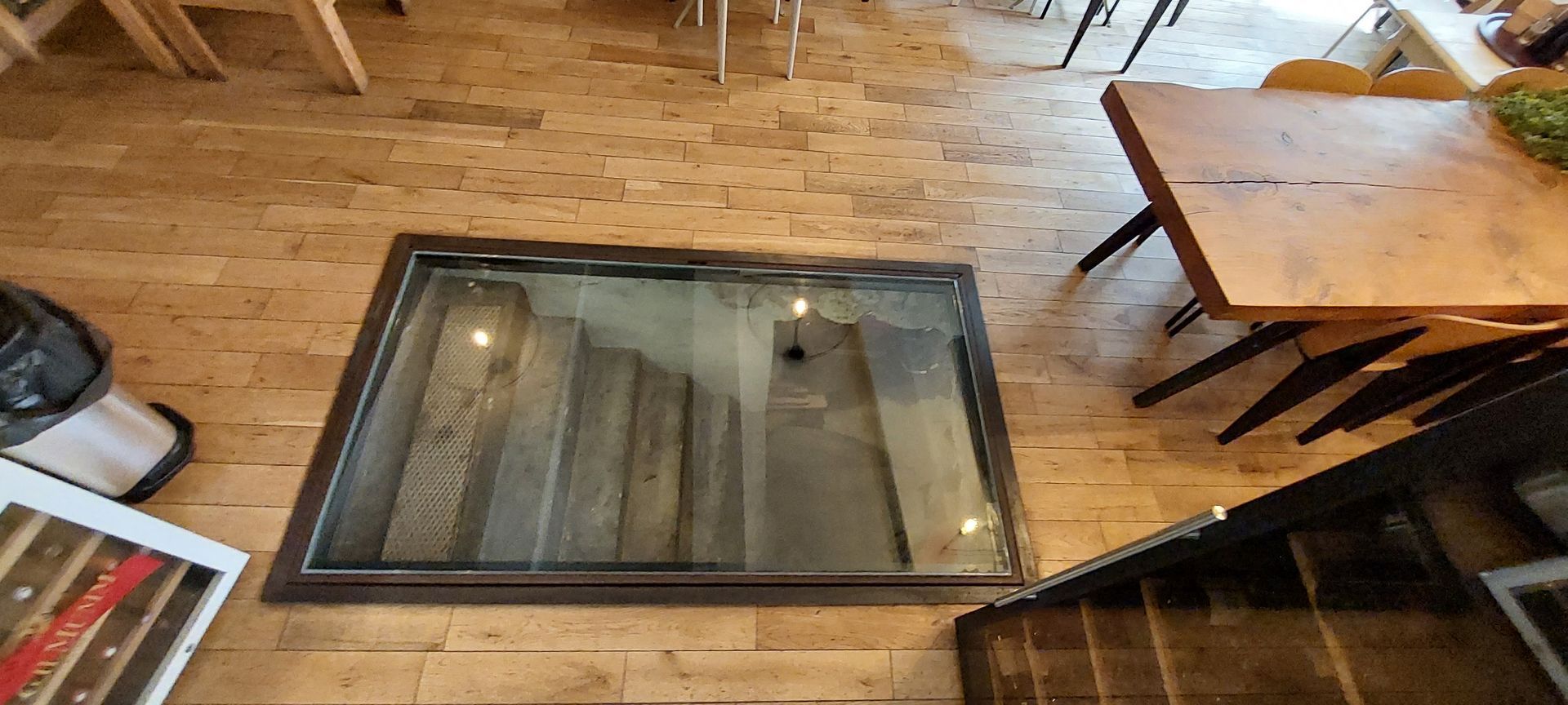Un sol en bois avec une fosse vitrée laissant apparaître un escalier, une table et des chaises.