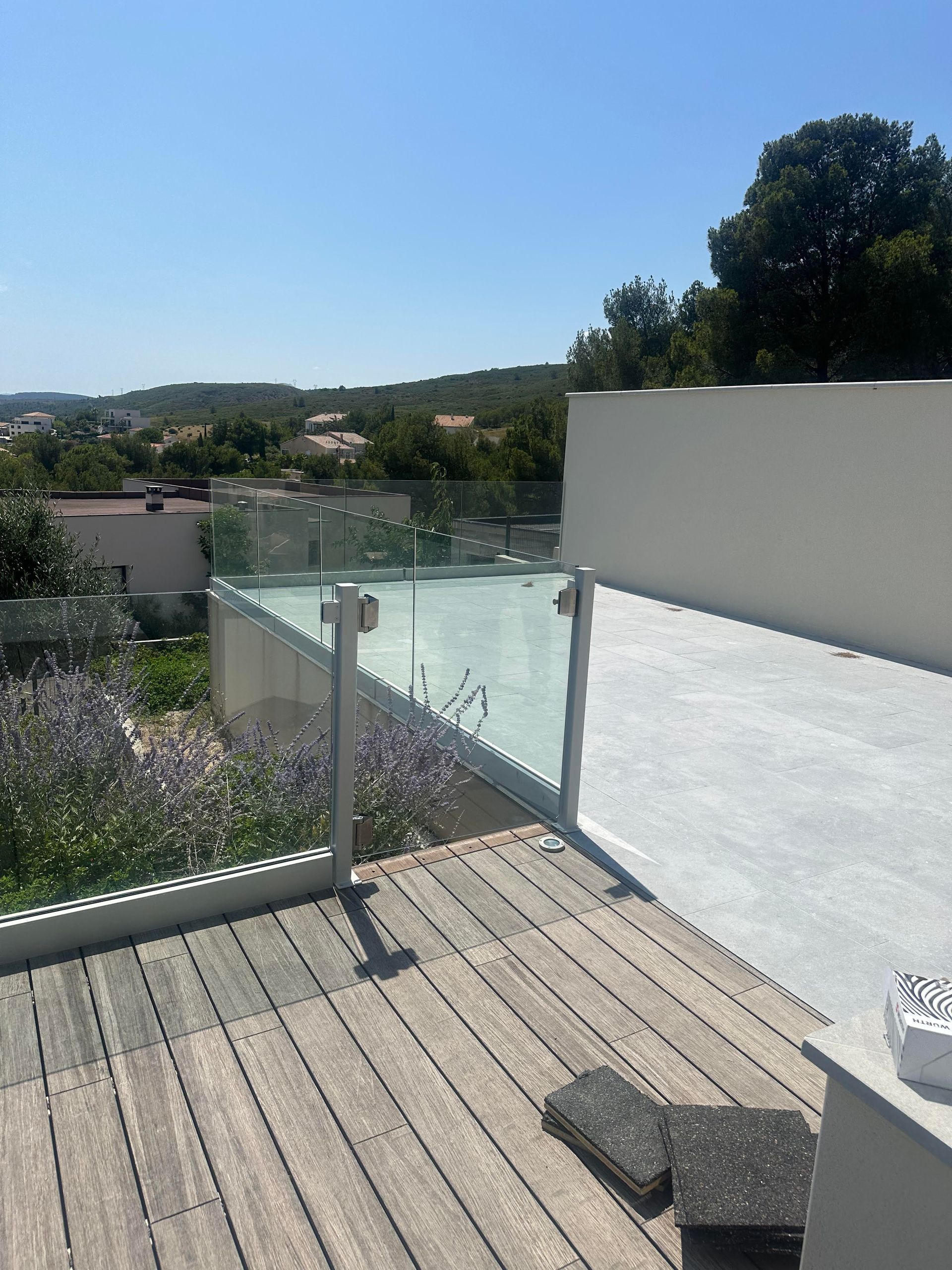 Terrasse sur le toit avec garde-corps en verre. Terrasse en bois, mur blanc, ciel bleu et paysage verdoyant au loin.