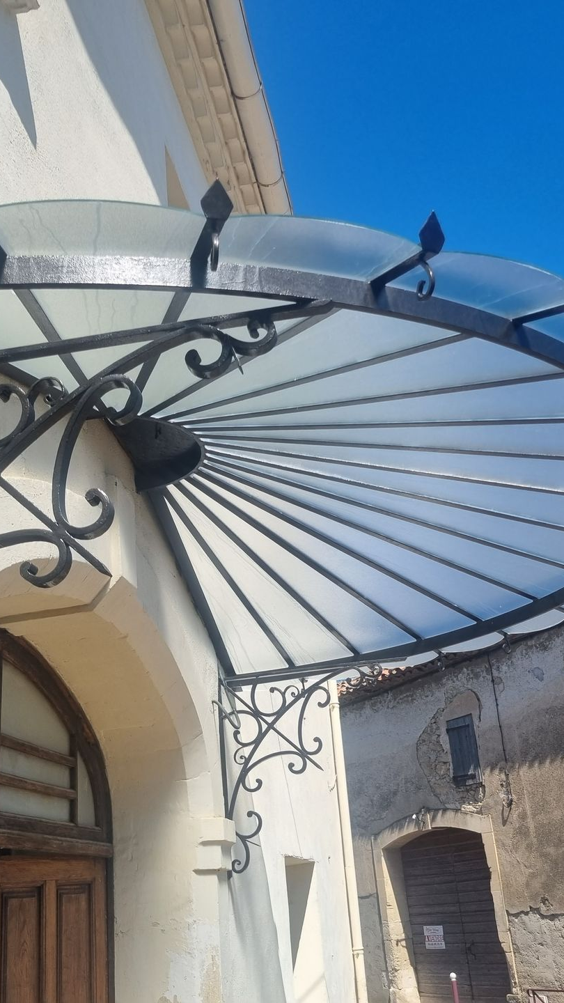 Auvent en fer forgé noir avec toit en verre au-dessus de l'entrée d'un bâtiment. Ciel bleu en arrière-plan.