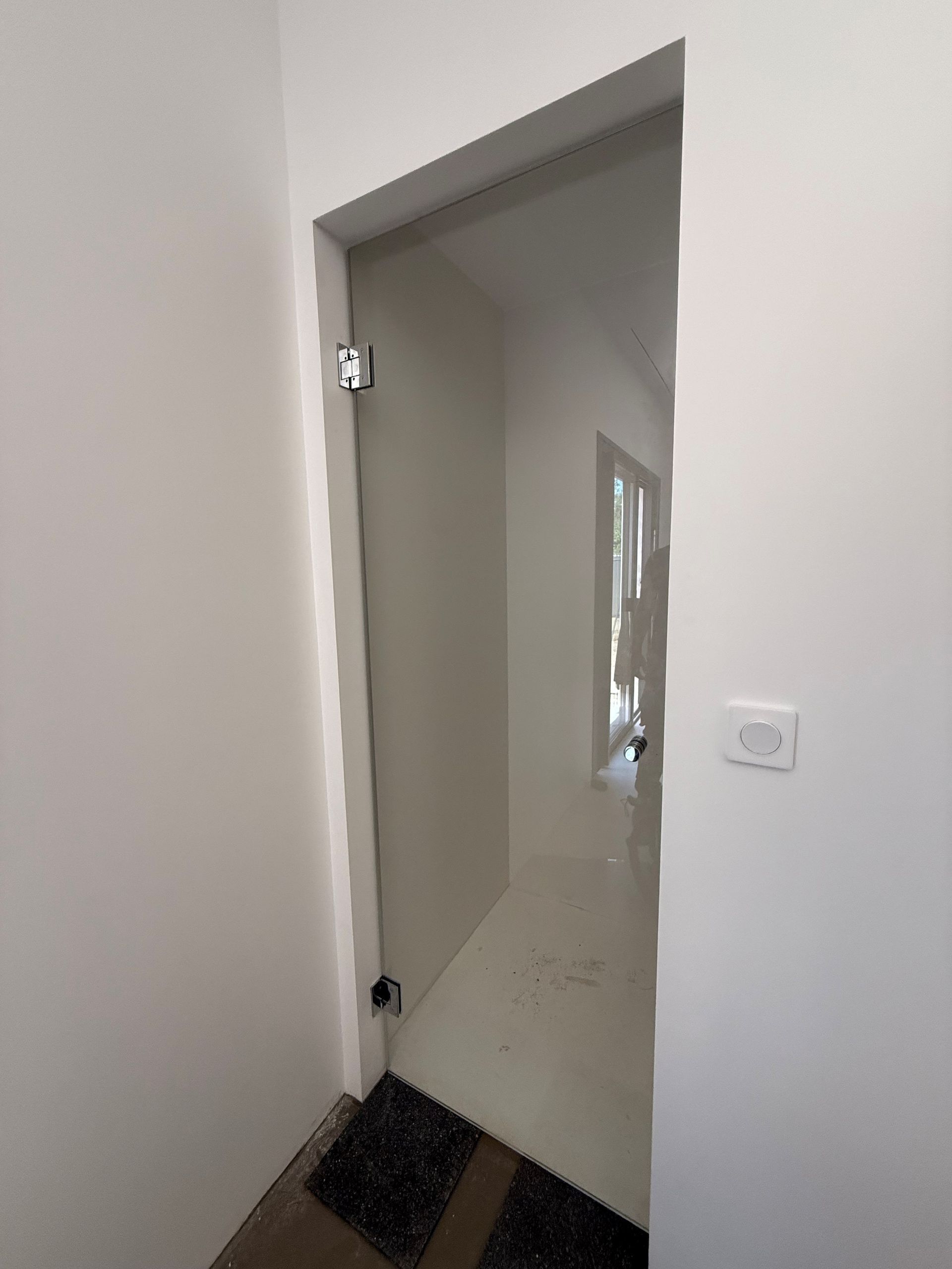 Porte vitrée dans un cadre blanc, donnant sur un couloir blanc où l'on aperçoit un interrupteur.