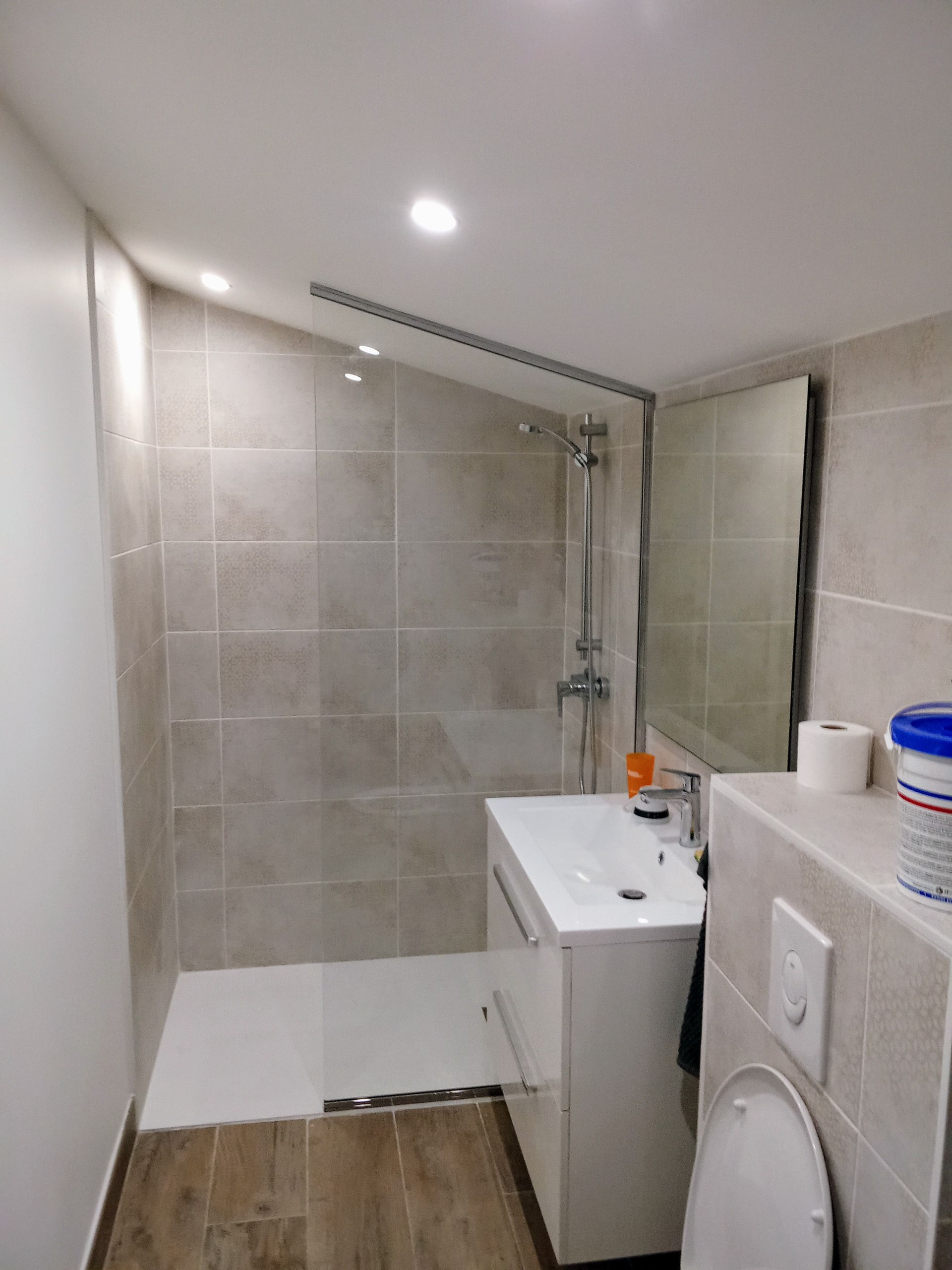 Petite salle de bains carrelée avec douche, lavabo, toilettes et miroir. Tons blancs et beiges dominants.