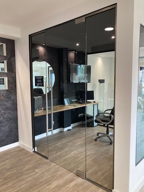 Porte de bureau vitrée avec cadre noir reflétant un intérieur de bureau : bureau, chaise et ordinateur.