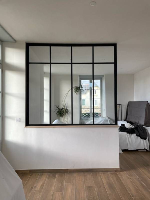Une cloison vitrée à cadre noir sépare deux pièces. Murs blancs, parquet. Vue sur l'extérieur par une fenêtre.