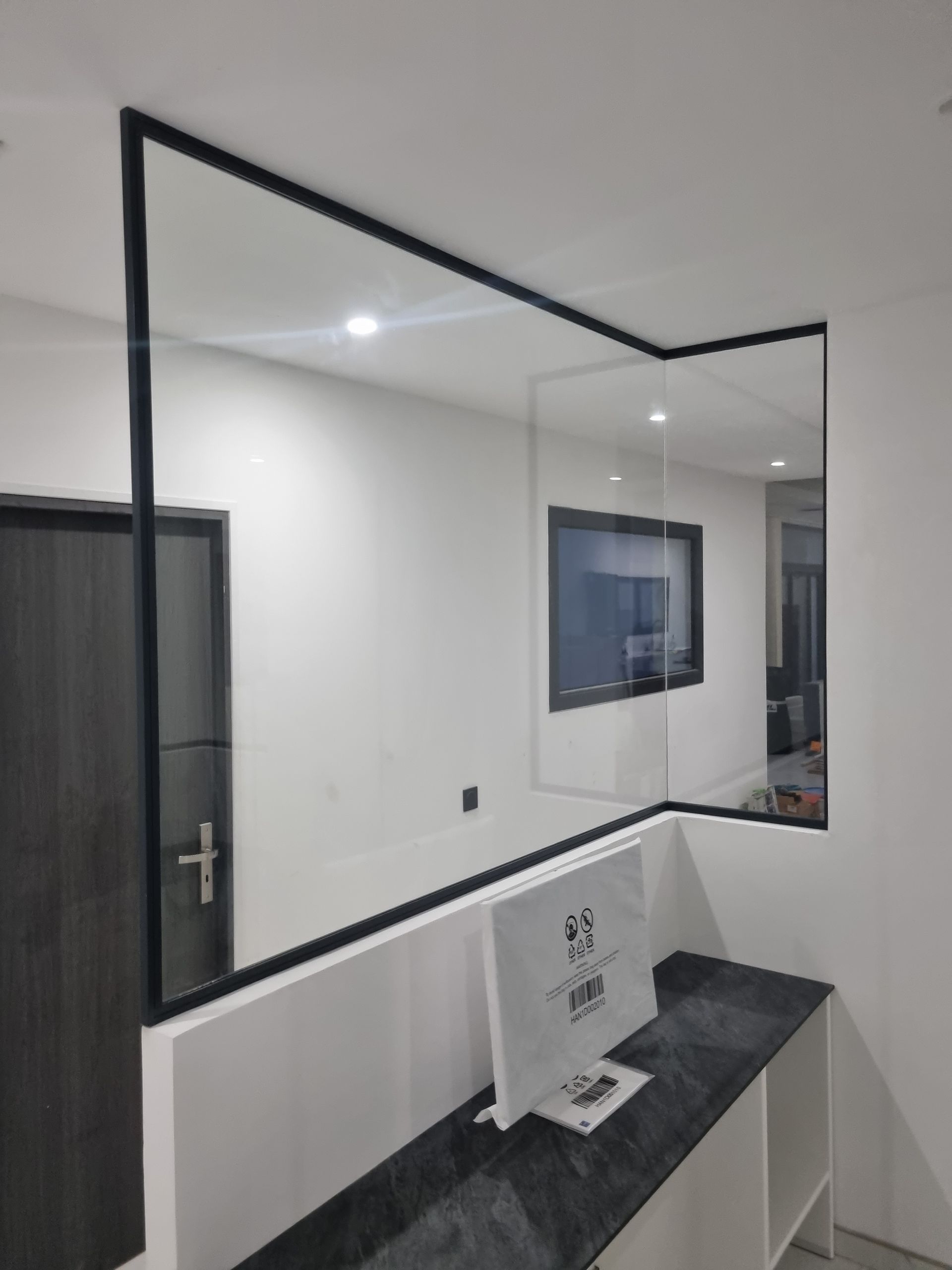 Cloison vitrée à cadre noir avec comptoir et télévision visibles dans un espace de bureau moderne.