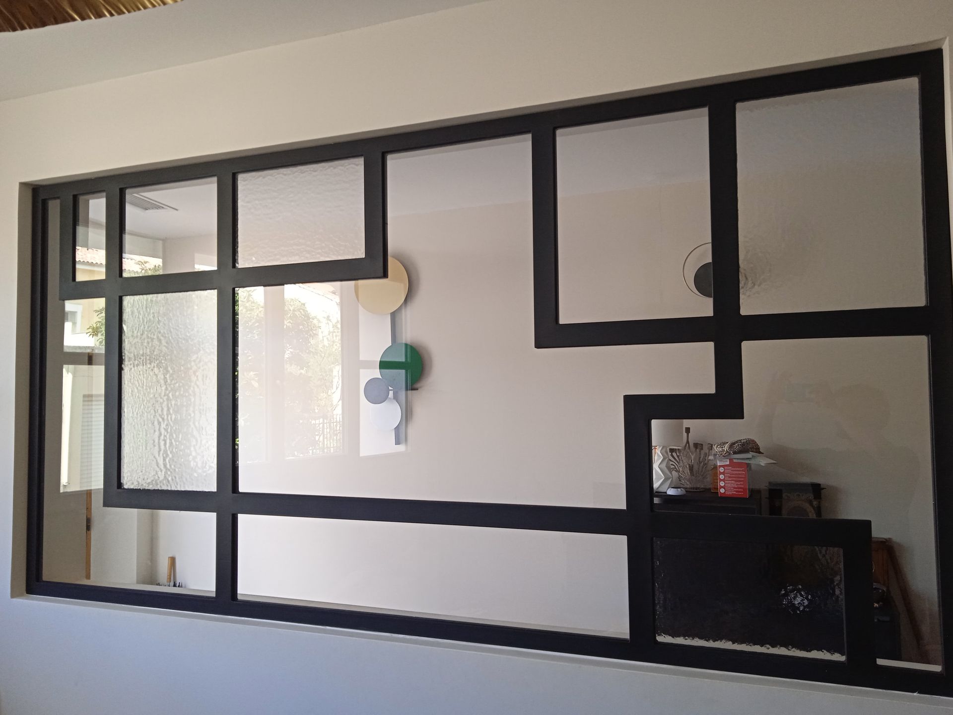 Cloison vitrée à cadre noir séparant deux pièces. Vitrages dépolis et transparents, murs blancs, décor minimaliste visible à travers les ouvertures.