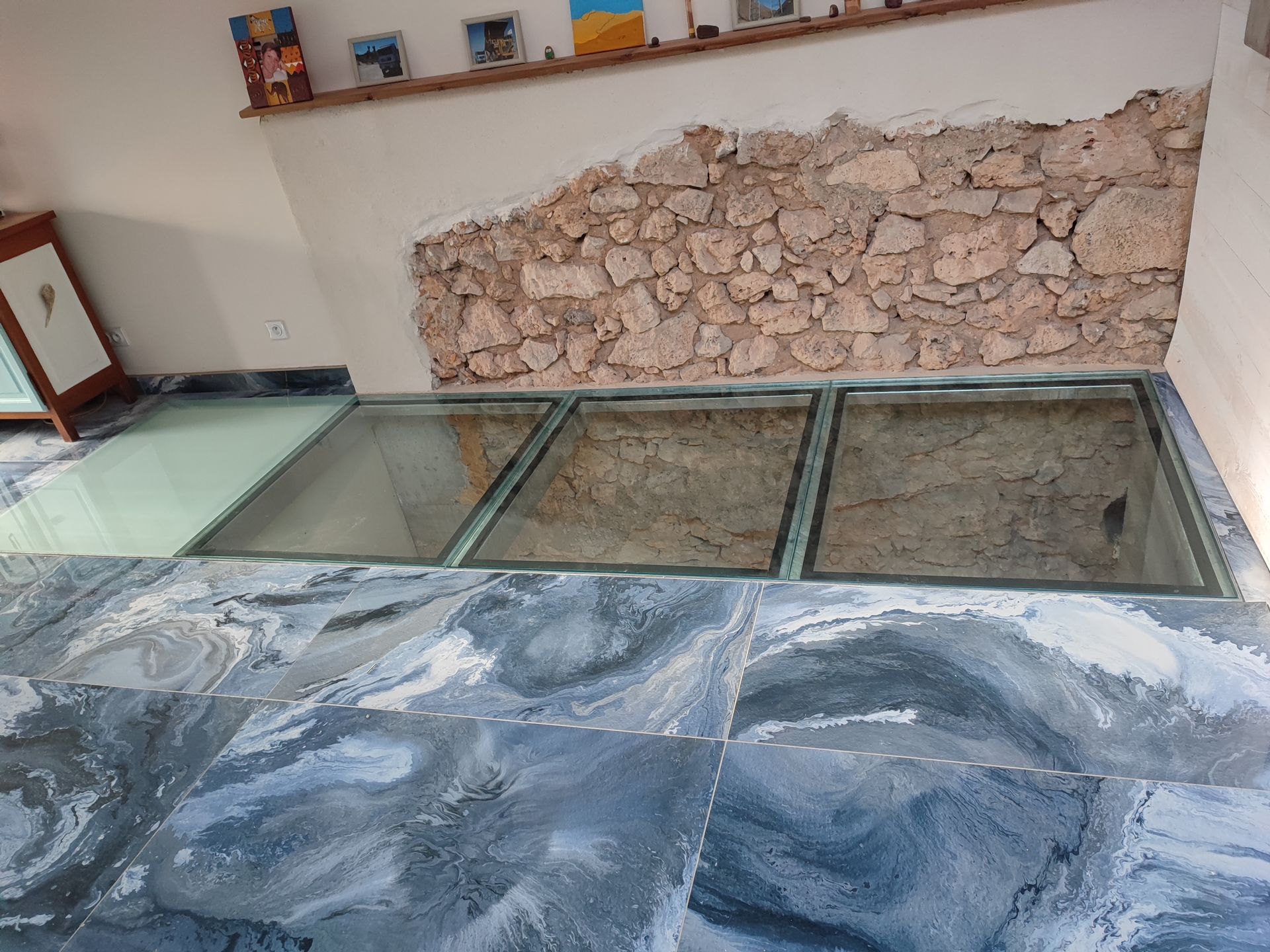 Des panneaux de verre au sol laissent entrevoir la pierre et la terre en dessous, dans une pièce avec un mur en pierre et un sol en carrelage bleu à motifs.