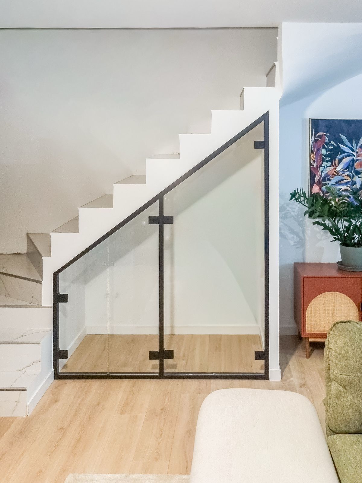Escalier avec rampe en verre, cadre noir et rangements en dessous. Parquet clair, murs blancs et tableau coloré.