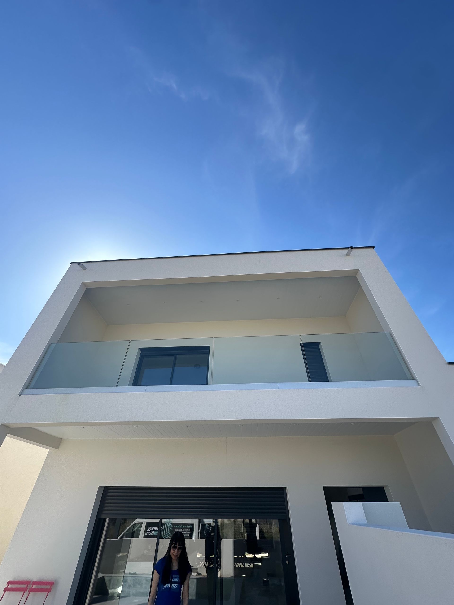 Immeuble blanc de deux étages avec un balcon vitré, sous un ciel d'un bleu éclatant. Une personne se tient près de l'entrée.