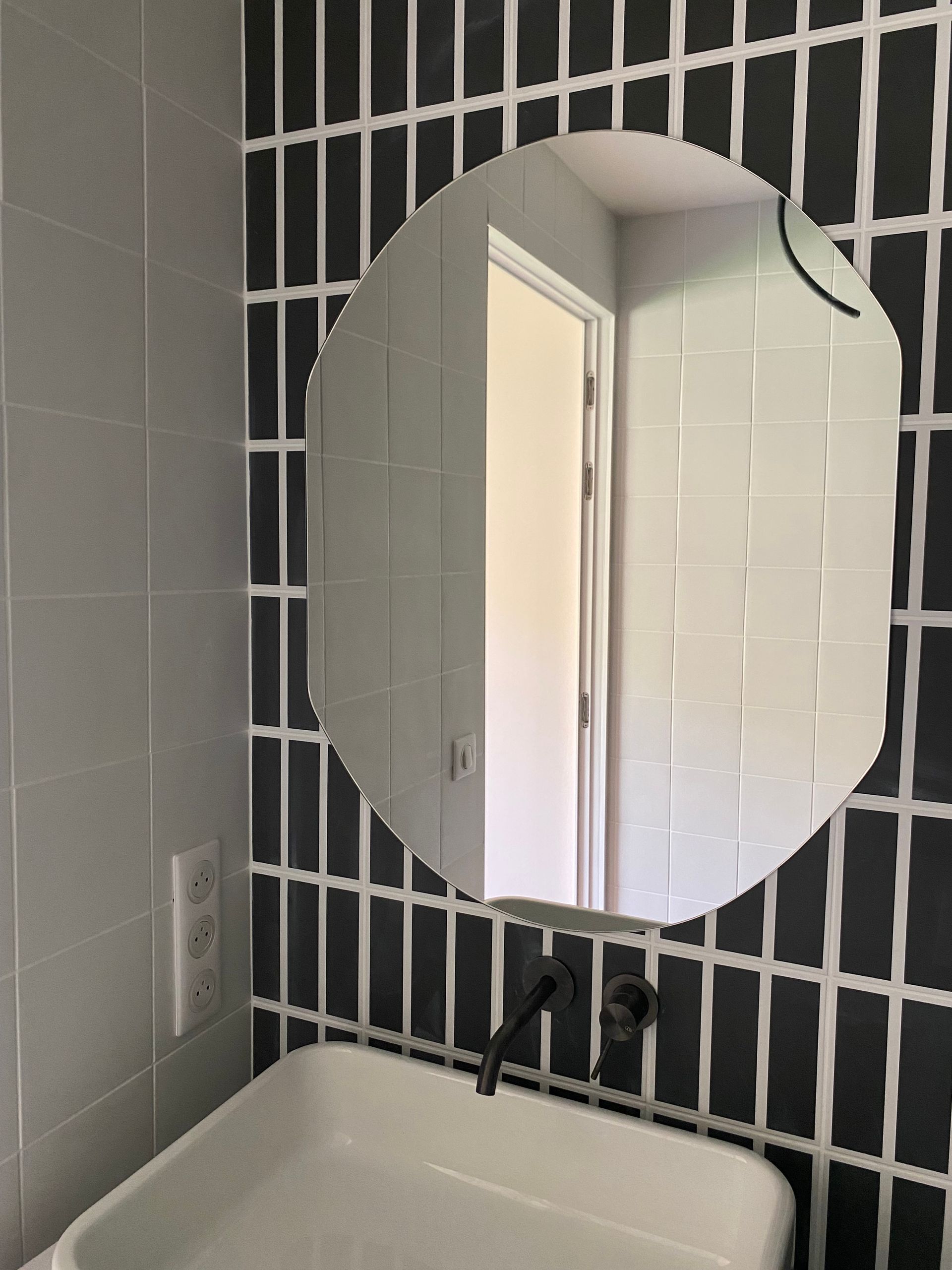 Salle de bains avec un miroir de forme irrégulière au-dessus d'un lavabo blanc, et des murs carrelés en noir et blanc.