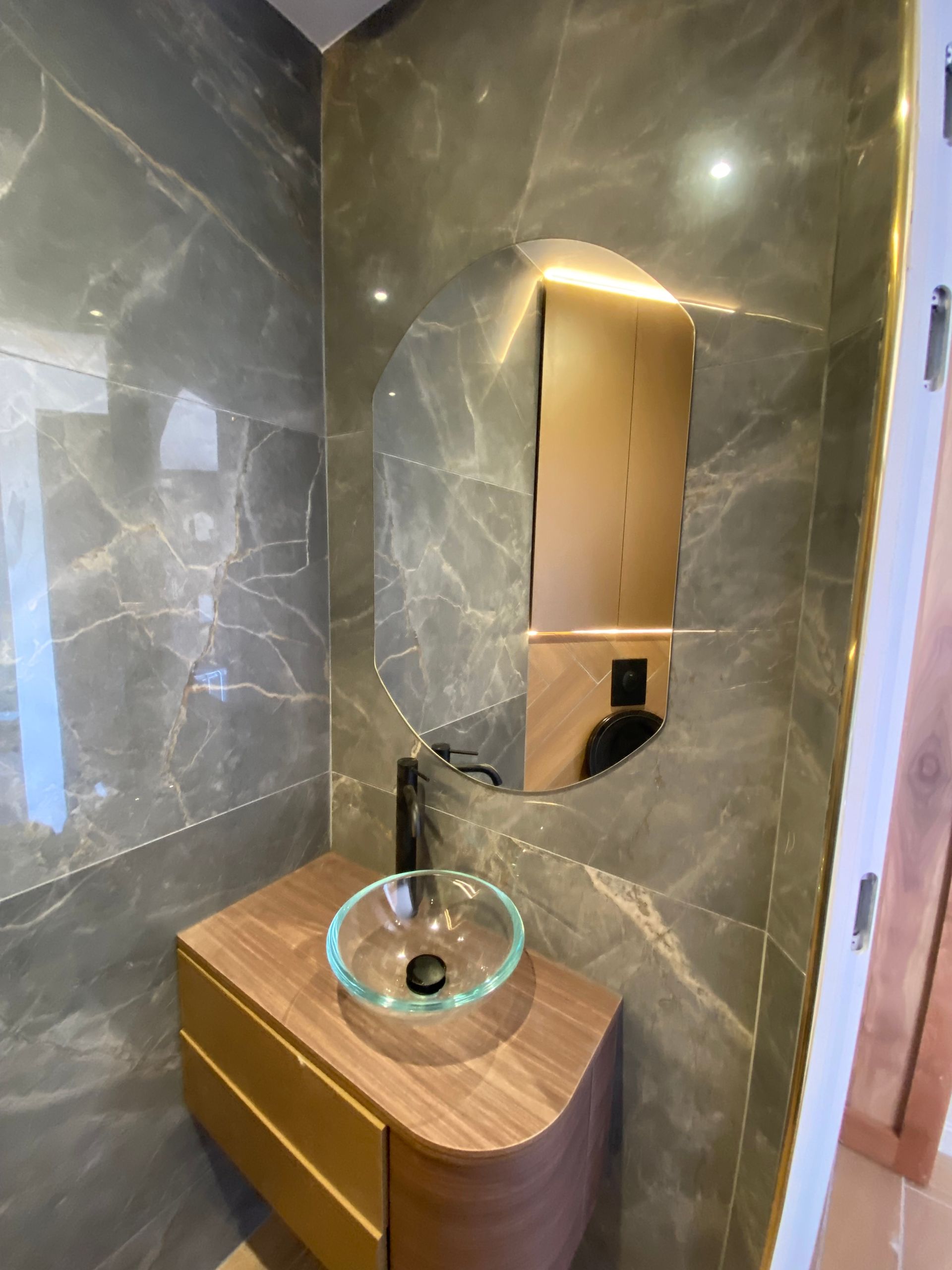 Salle de bains avec carrelage en marbre gris, meuble-lavabo en bois, vasque en verre et miroir rond.