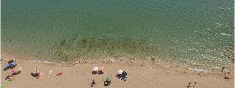 Vista aerea di una spiaggia con acqua turchese cristallina e persone che prendono il sole sotto gli ombrelloni.