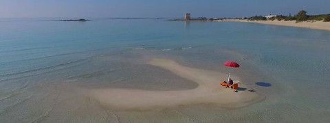 Una spiaggia con acqua cristallina color turchese. C'è una piccola isola di sabbia con un ombrellone rosso e due sedie.