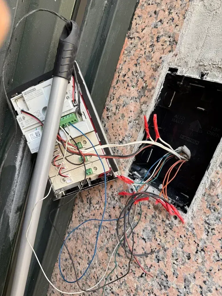 Caja eléctrica abierta con cables expuestos, placa de circuito y poste de metal cerca de un edificio.
