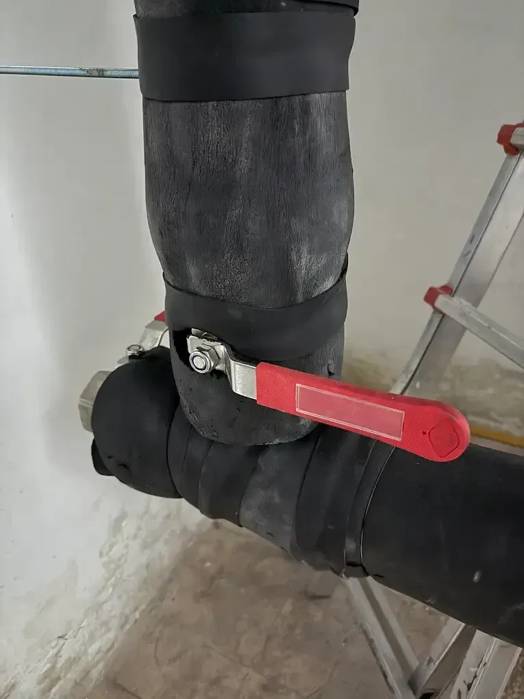 Tubos aislados de color negro con una válvula con manija roja, cerca de una escalera de mano, contra una pared blanca.