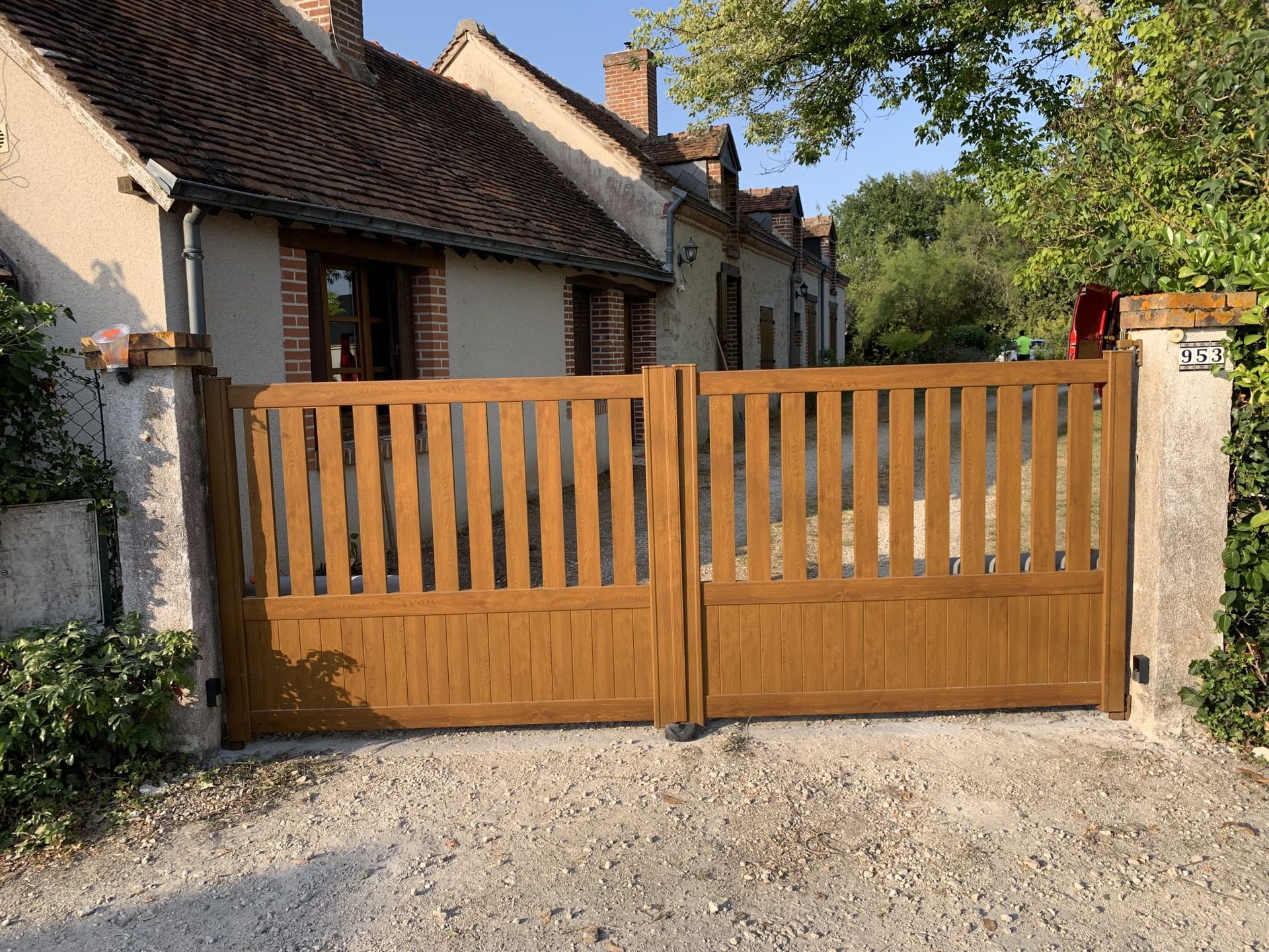 Portail en bois devant une maison de campagne, avec des piliers en pierre et une allée en gravier