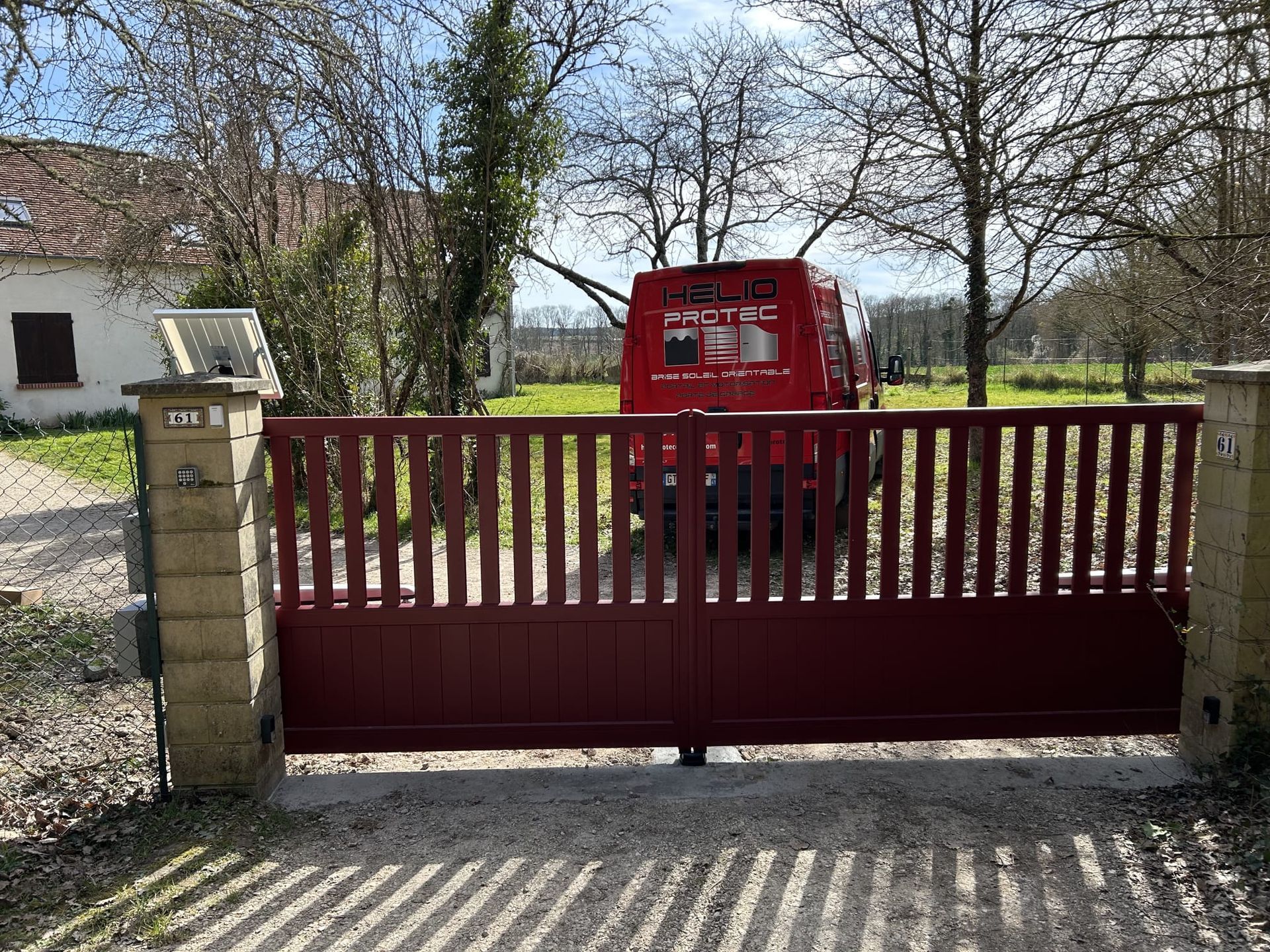 Portail rouge dans une cour à la campagne, avec un véhicule rouge derrière, des arbres et un petit bâtiment en arrière-plan.
