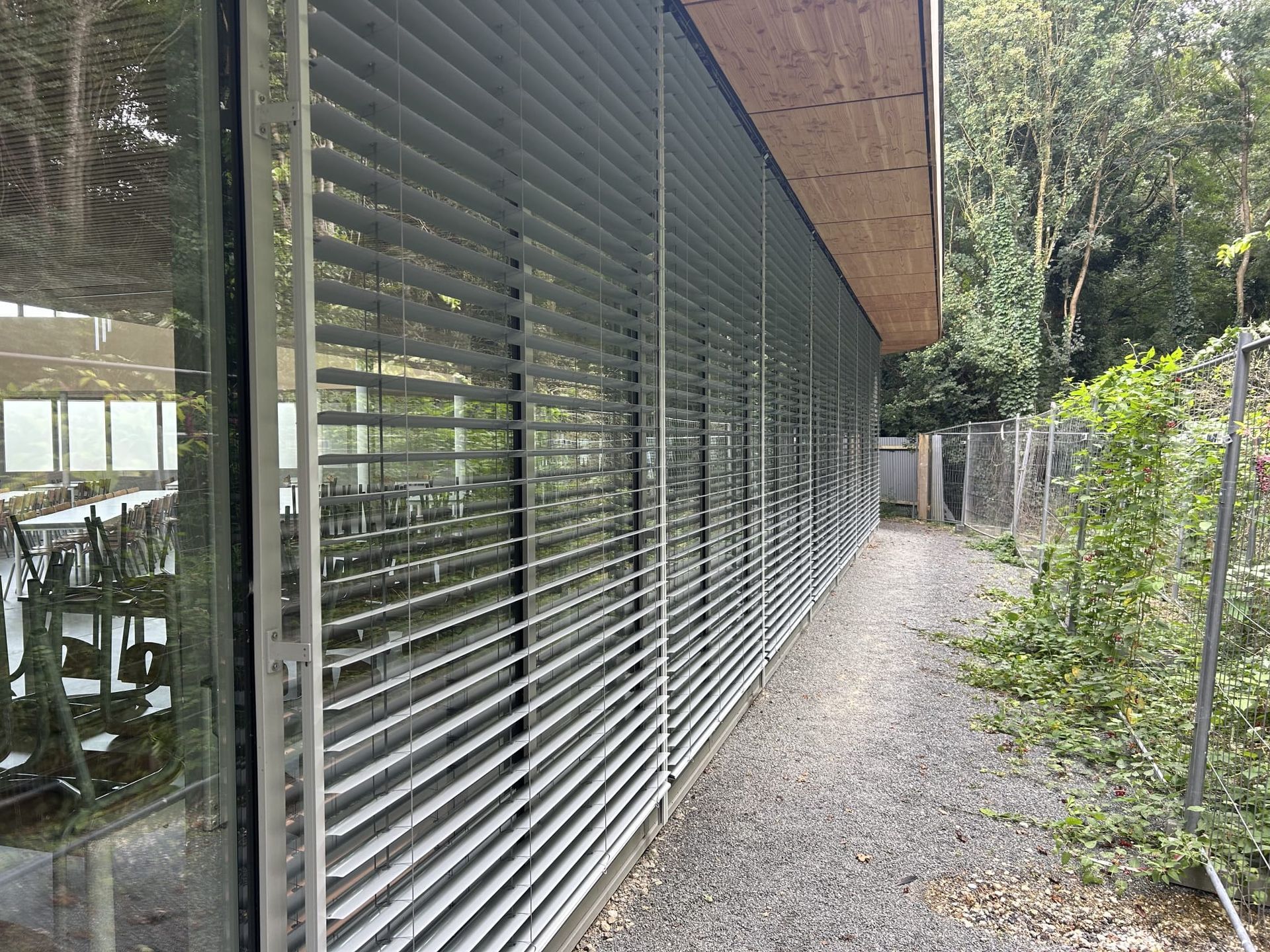 Longue paroi vitrée avec des stores horizontaux, le long d’un chemin de gravier bordé de verdure