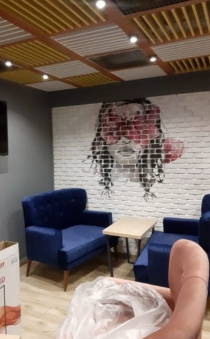 Pièce avec fauteuils bleus, petite table et fresque murale en briques représentant le visage d'une femme.