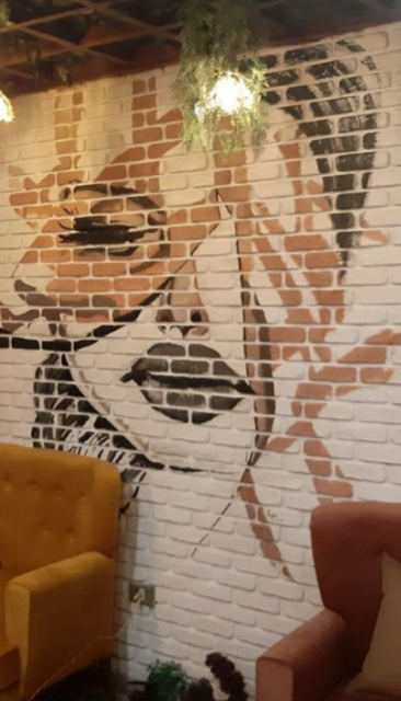 Fresque représentant le visage d'une femme sur un mur de briques, avec des chaises jaunes et rouges au premier plan.