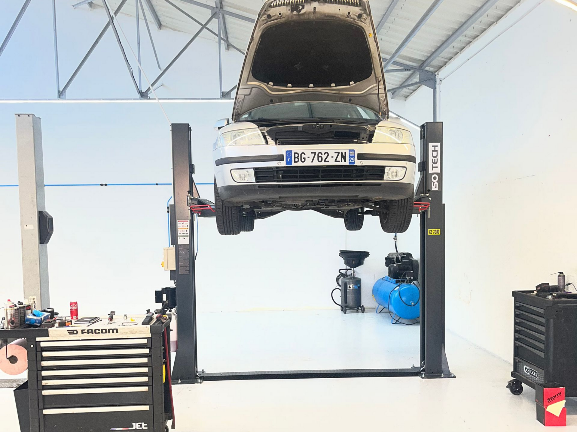 Une voiture au capot ouvert, soulevée par un pont élévateur hydraulique à deux colonnes.